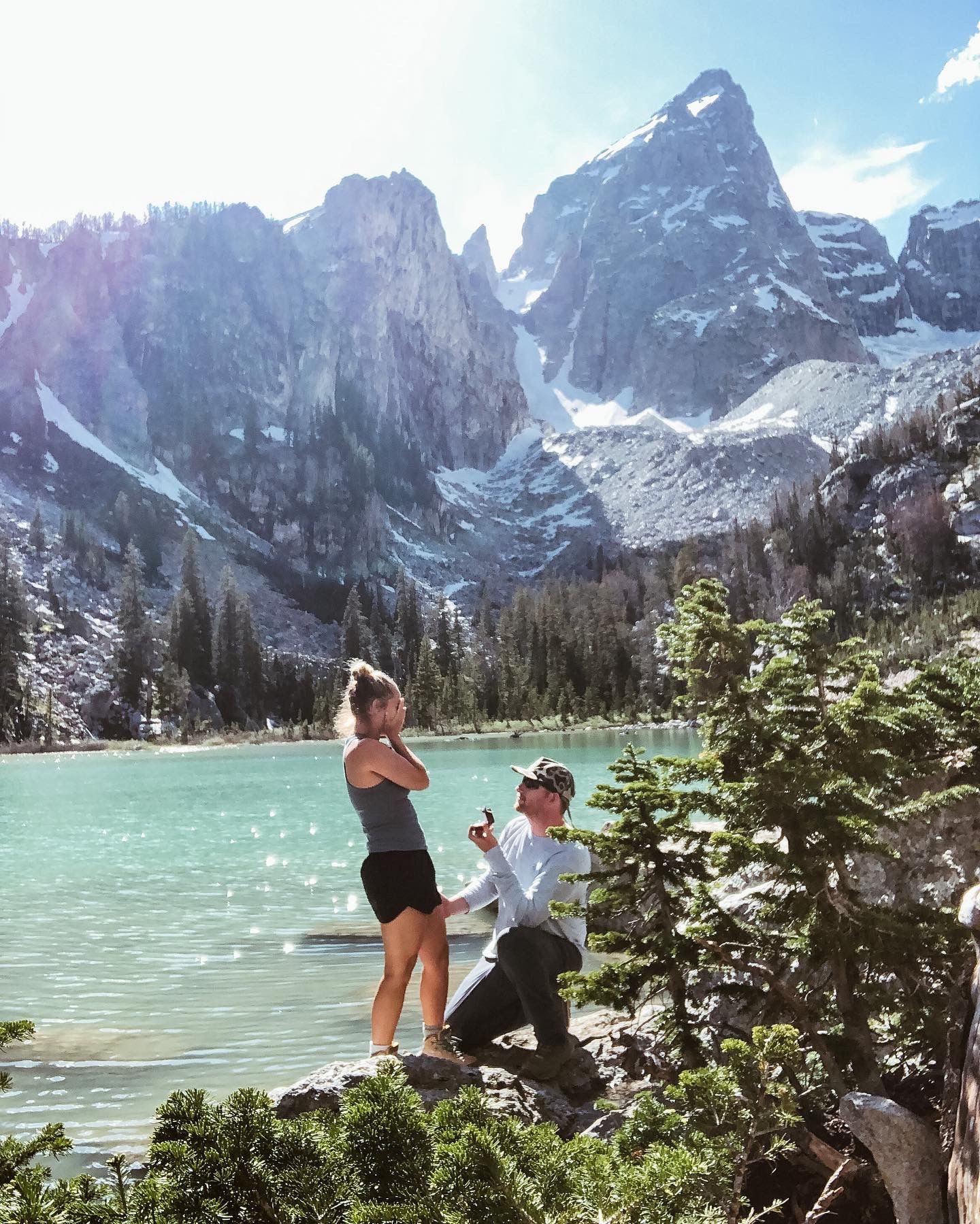 Man proposes to woman on rocky ledge with stunning turquoise alpine lake and Teton mountains behind.