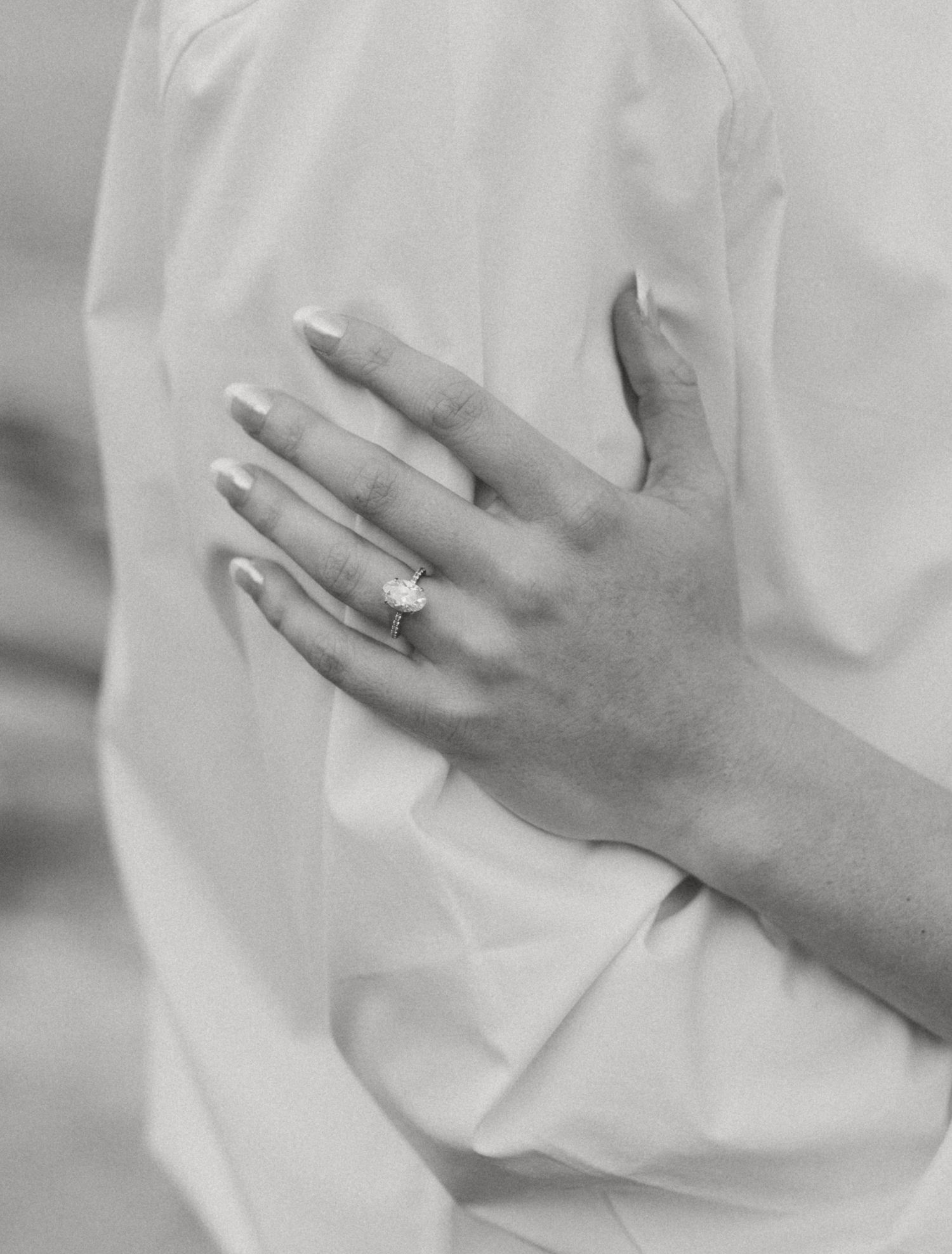 Black and white close-up of woman wearing oval diamond engagement ring on white shirt.