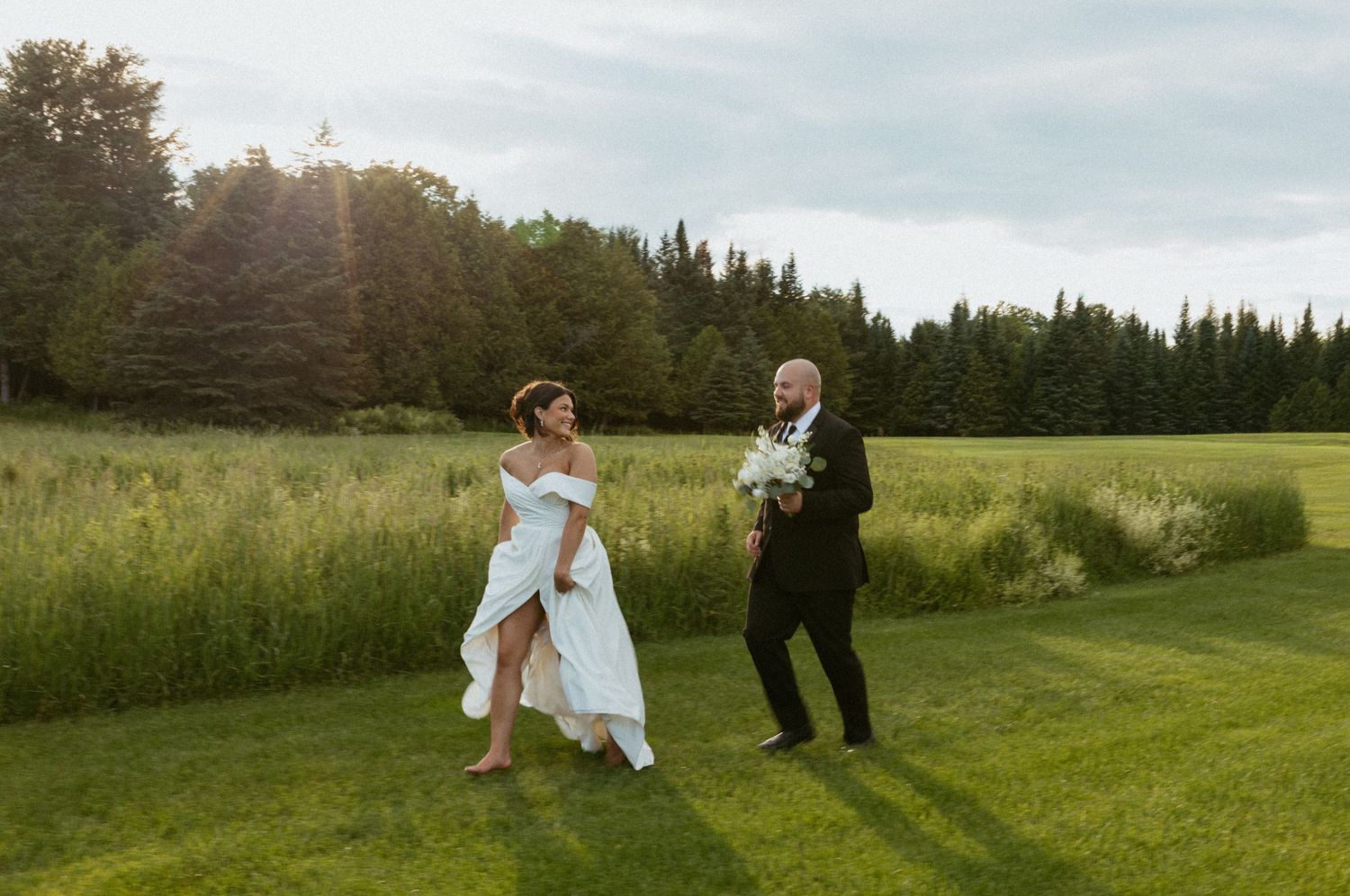 Bride and groom walking across lush green lawn at golden hour, with tall trees in the background.