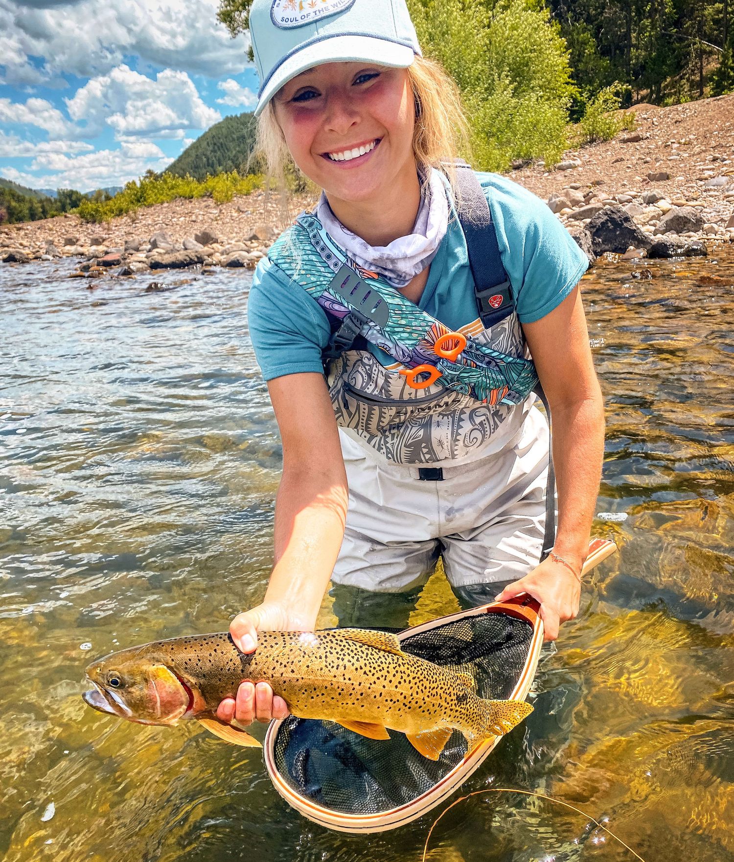 Smiling blonde woman in waders proudly holding large brown trout caught fly fishing in mountain stream.