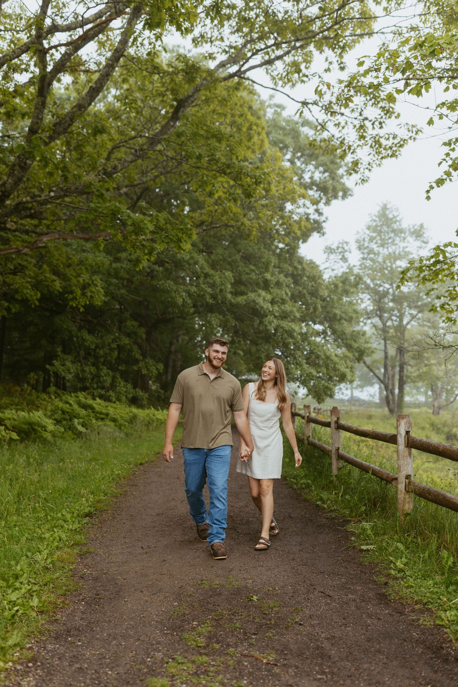 Happy couple walking hand in hand along a tree-lined Acadia path beside a rustic wooden fence.