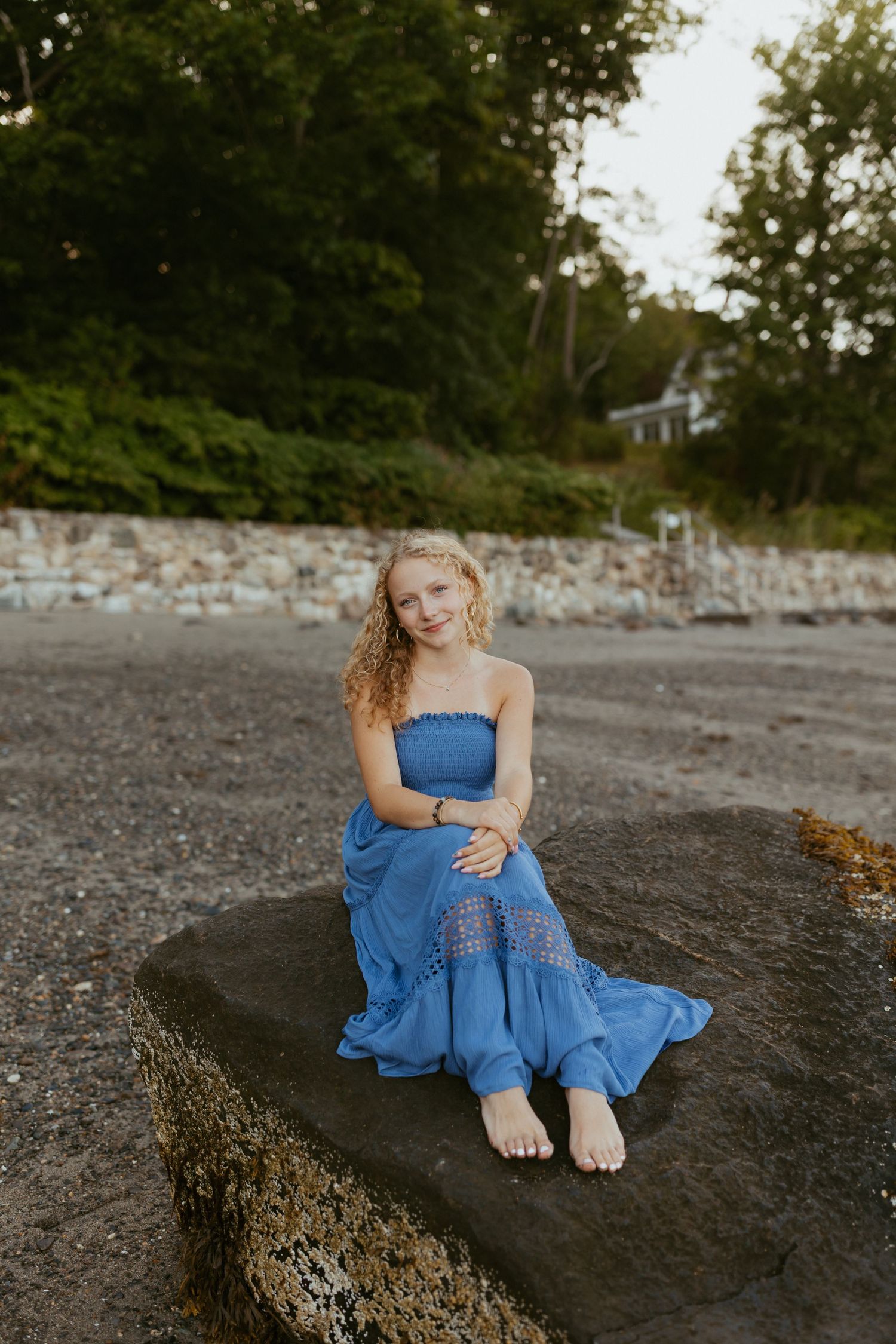 Young woman in blue strapless maxi dress sitting barefoot on a rock at a beach with green trees behind her.