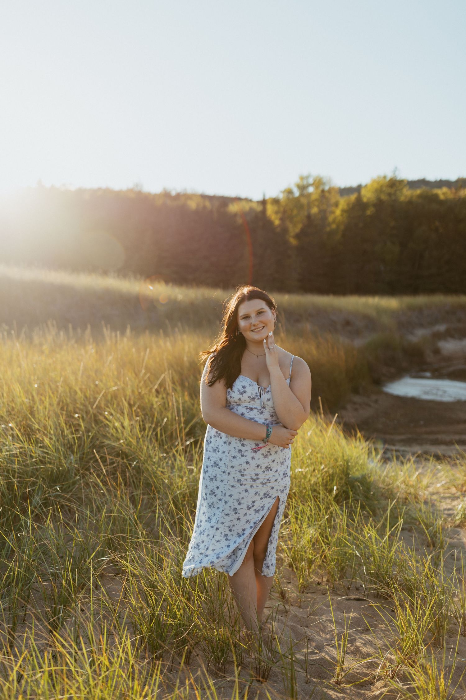 Woman in floral sundress smiling on sandy dunes with golden hour sunlight and green grass backdrop.
