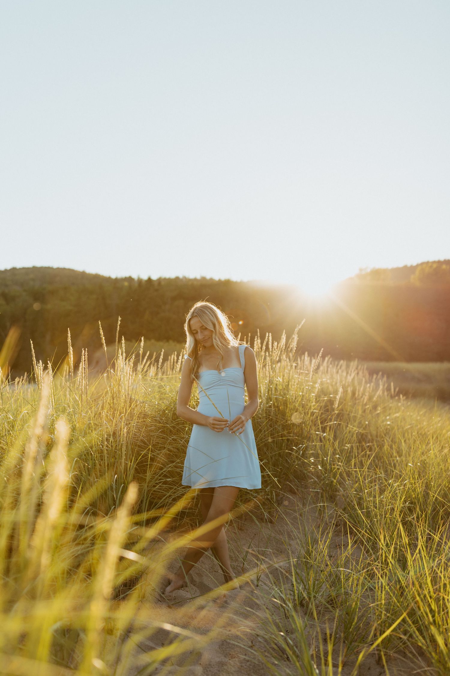 Pregnant woman in light blue dress standing in golden grass field at sunset, cradling baby bump.
