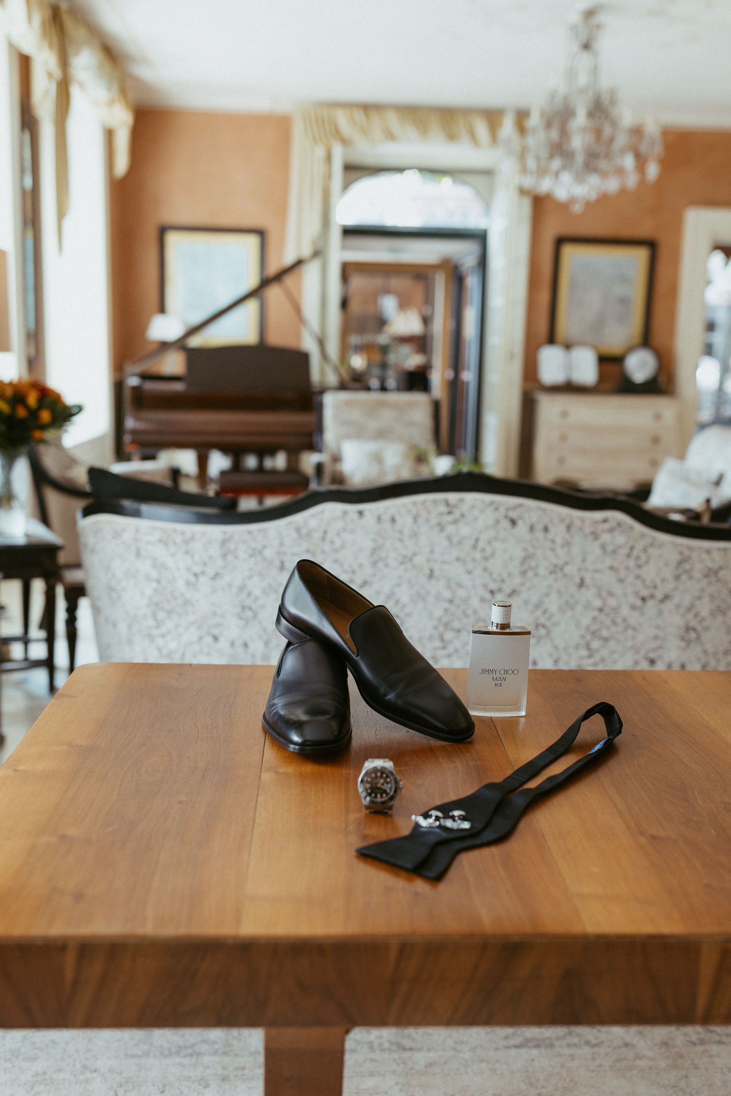 Black leather loafers, cologne, watch, and bow tie arranged on wooden table in elegant room with piano.
