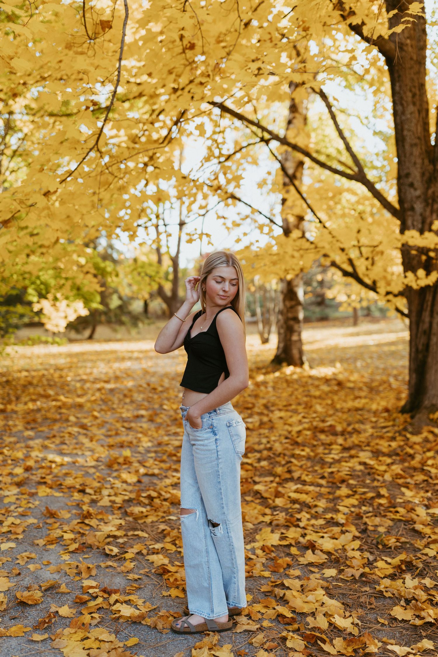 Young blonde woman in black crop top and light jeans posing in autumn park with golden yellow foliage.
