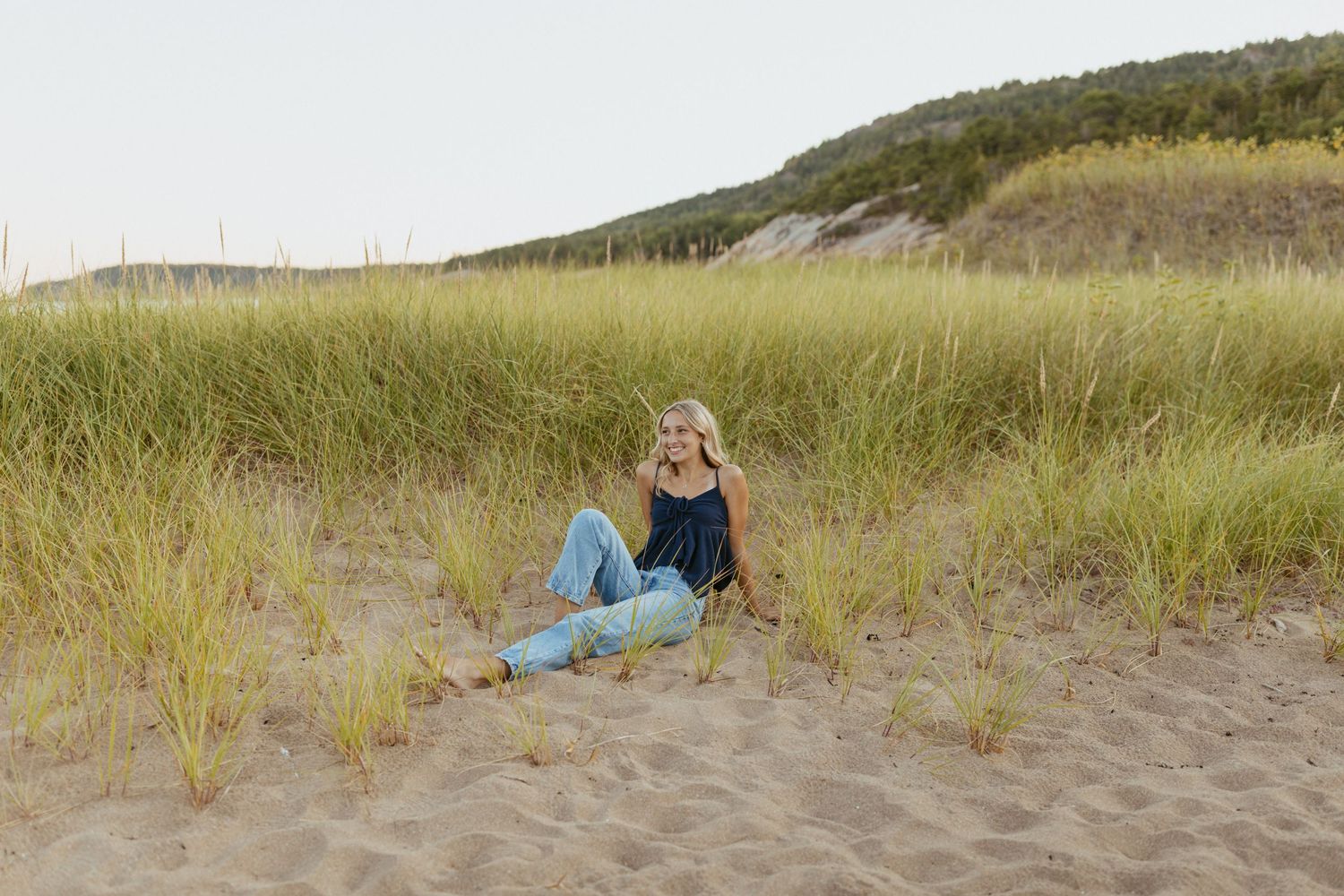 Blonde woman sitting among sand dunes and tall grass at a beach, wearing jeans and a dark top.