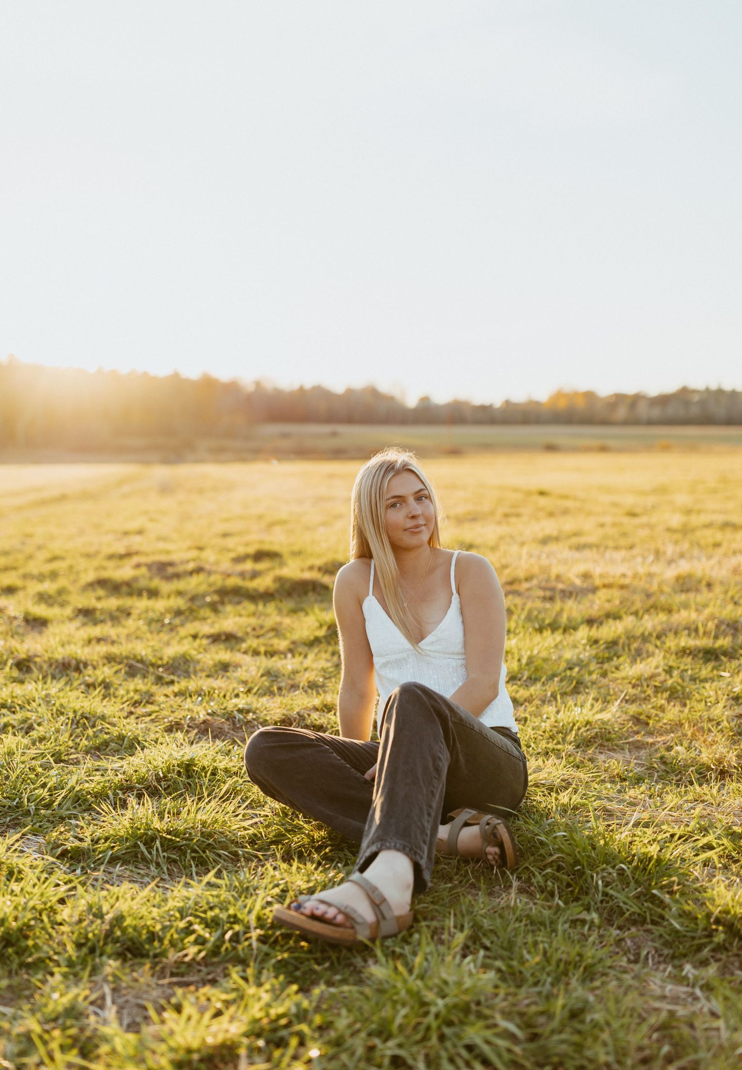 Blonde woman in white tank and dark jeans sits cross-legged on green grass field during golden hour sunset.