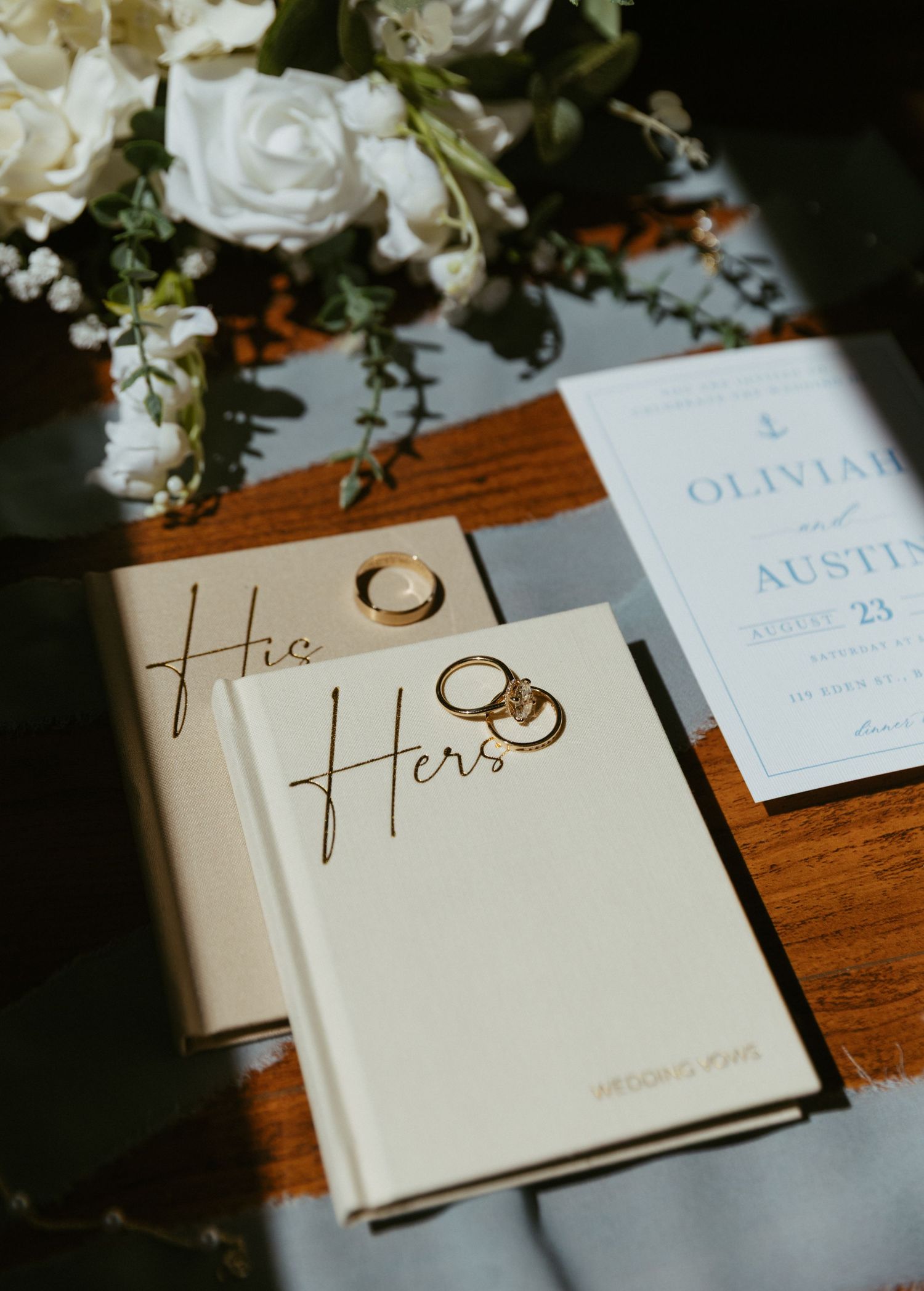 His and Hers wedding vow books with gold rings, white floral bouquet, and elegant wedding invitation on wooden table.