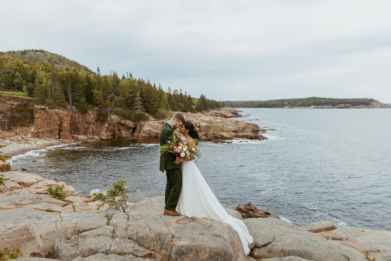 Bride and groom embrace on rocky coastal cliffs with evergreen trees and ocean views on their wedding day.