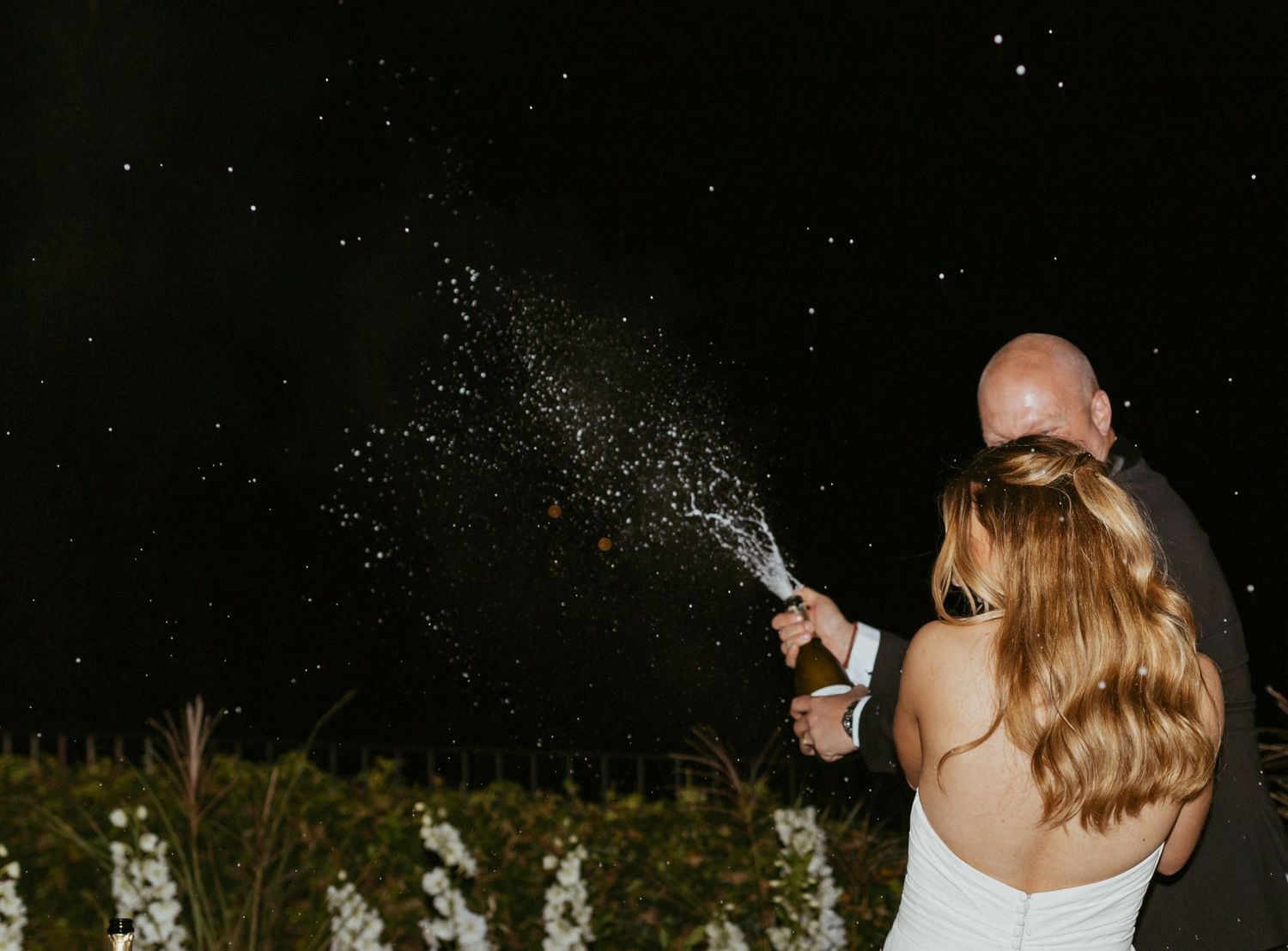 Bride and groom spraying champagne at night during wedding celebration outdoors.