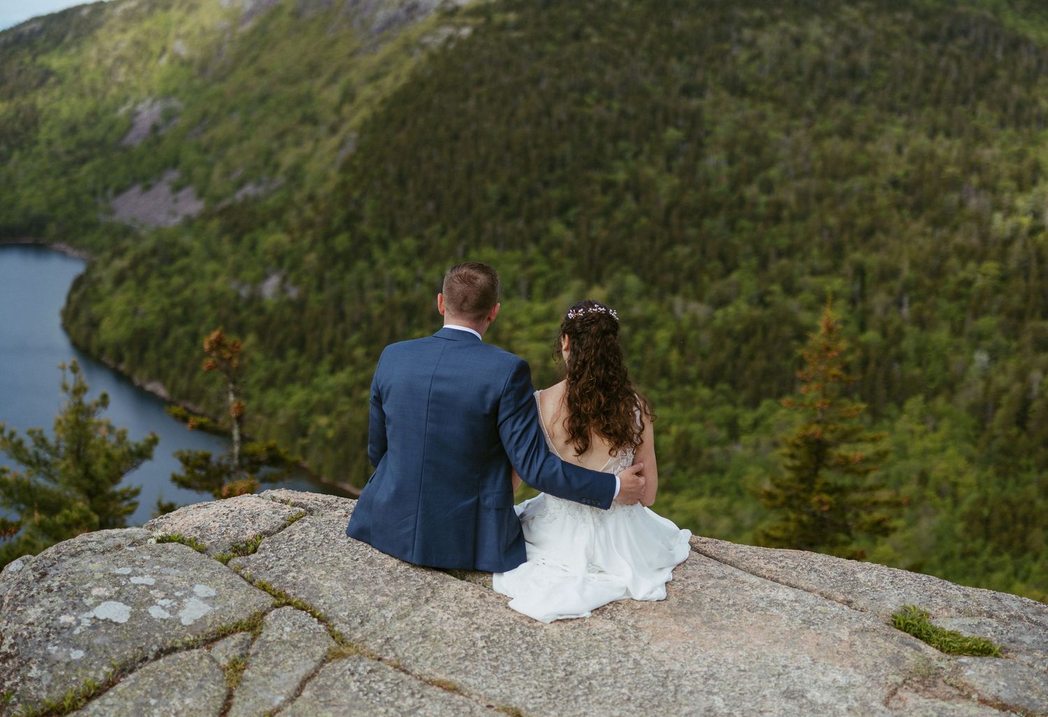 Bride and groom sitting on rocky mountain summit overlooking scenic lake and forest landscape during outdoor wedding.