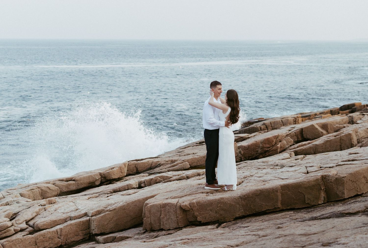 Couple holding hands on rocky coastal cliffs with dramatic ocean waves crashing behind them.