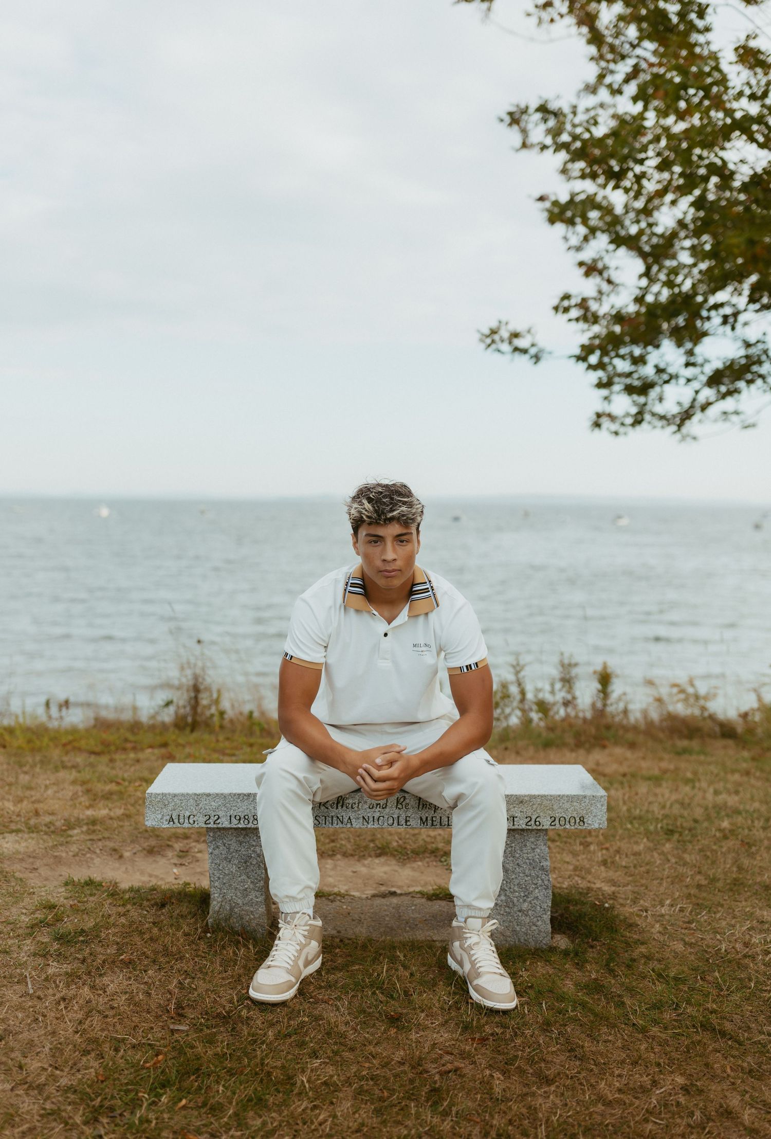 Young man in white polo shirt sitting on a memorial bench overlooking the ocean on a cloudy day.