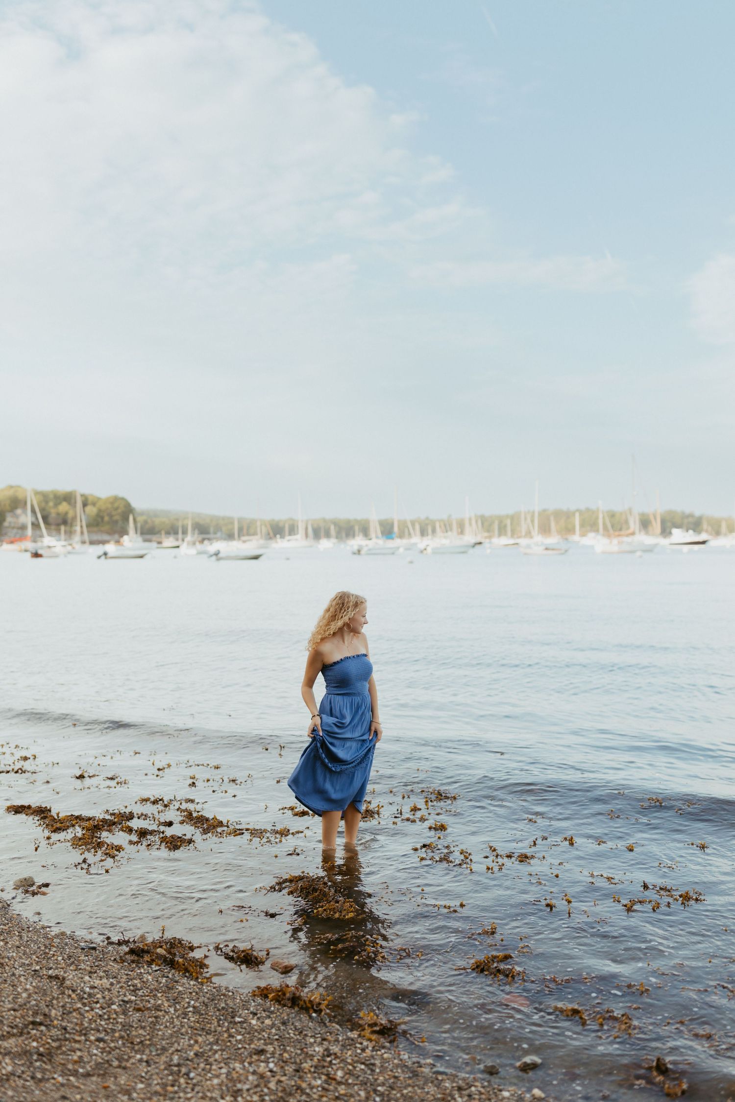 Woman in blue strapless dress standing at waters edge on rocky shore with sailboats in distance.