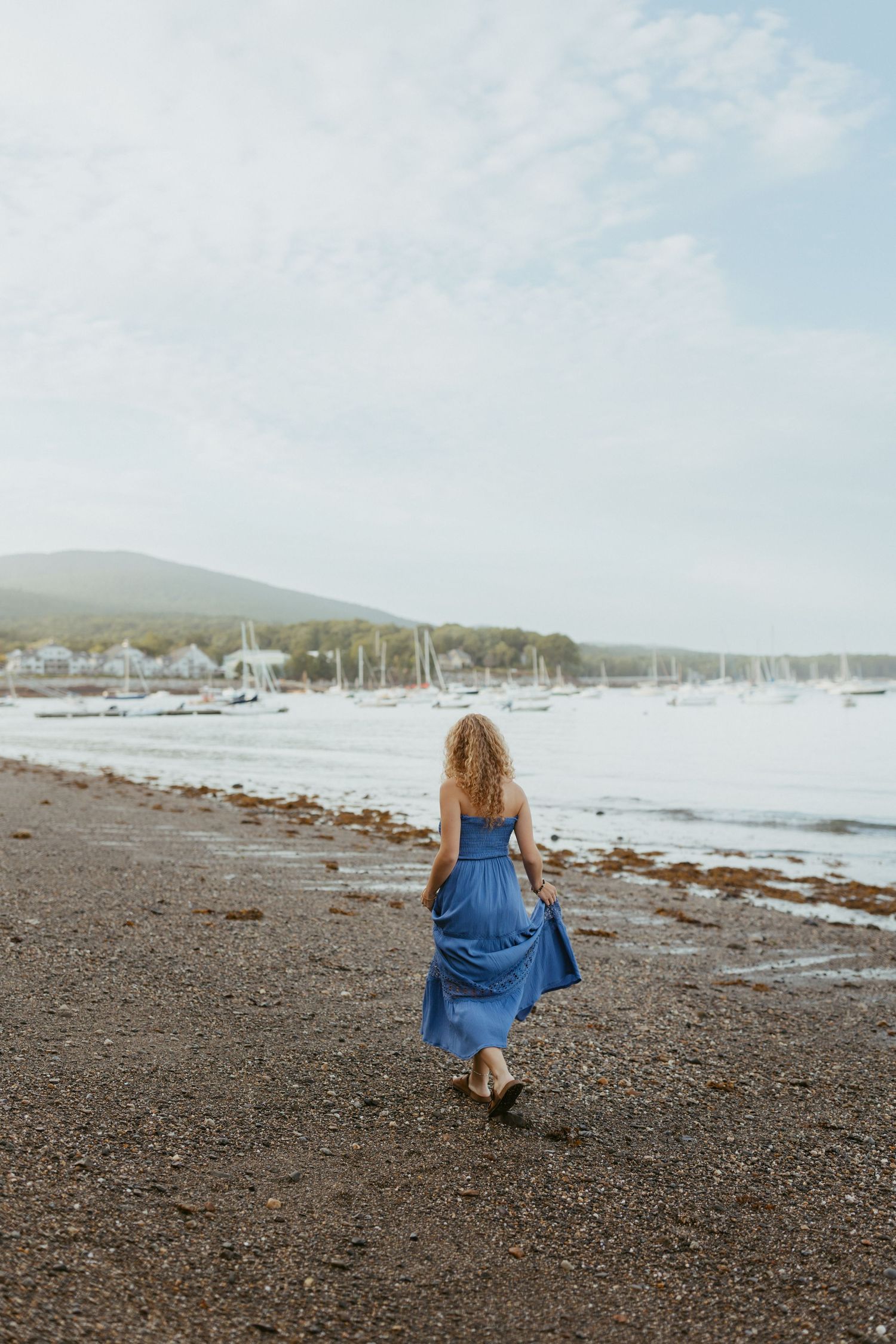 Woman in blue maxi dress walking along a rocky beach with sailboats and mountains in the background.