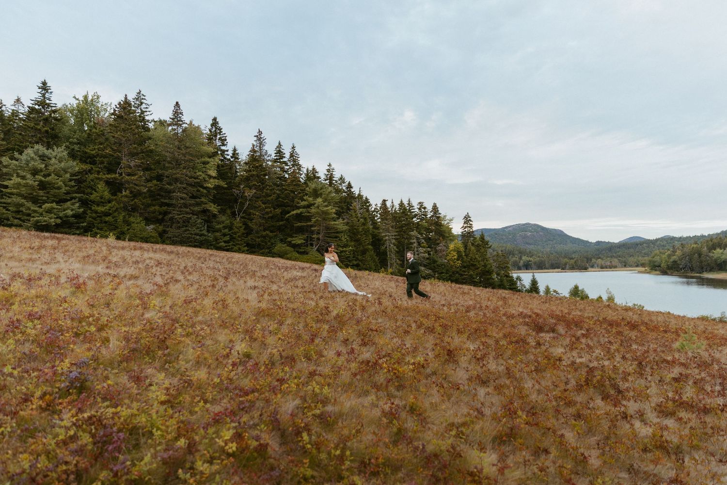 Bride and groom walk through an autumn meadow with a lake and forest backdrop.