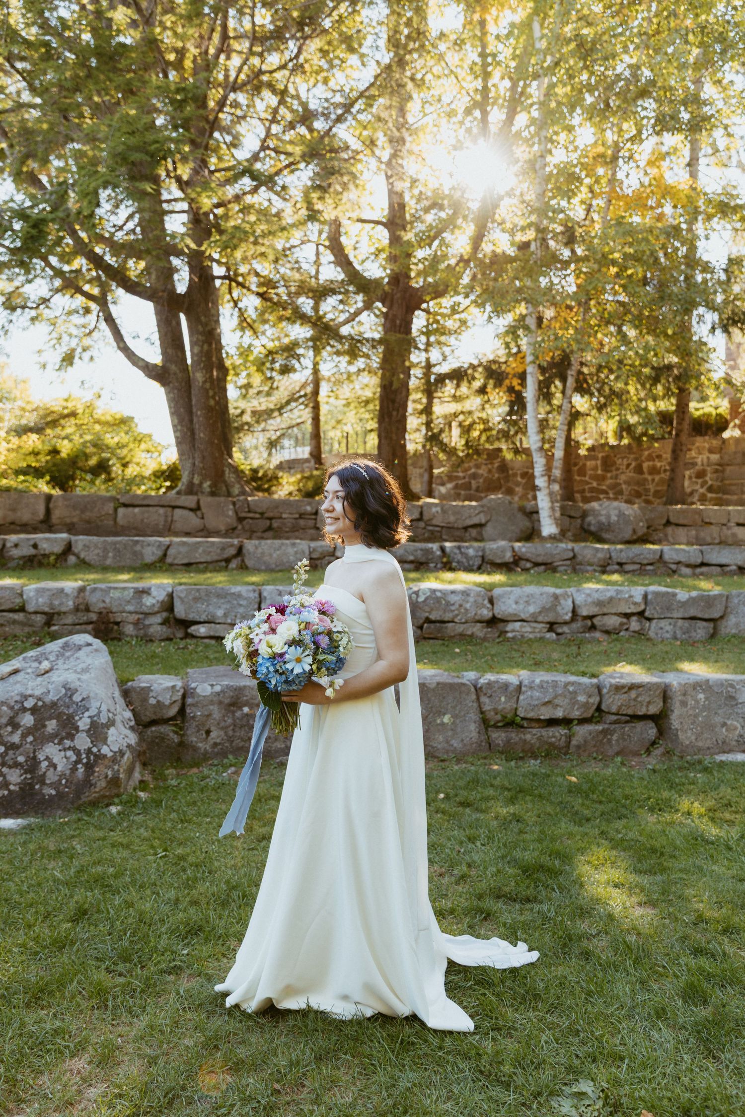 Bride in white gown holds a wildflower bouquet in a sunlit garden with stone walls.