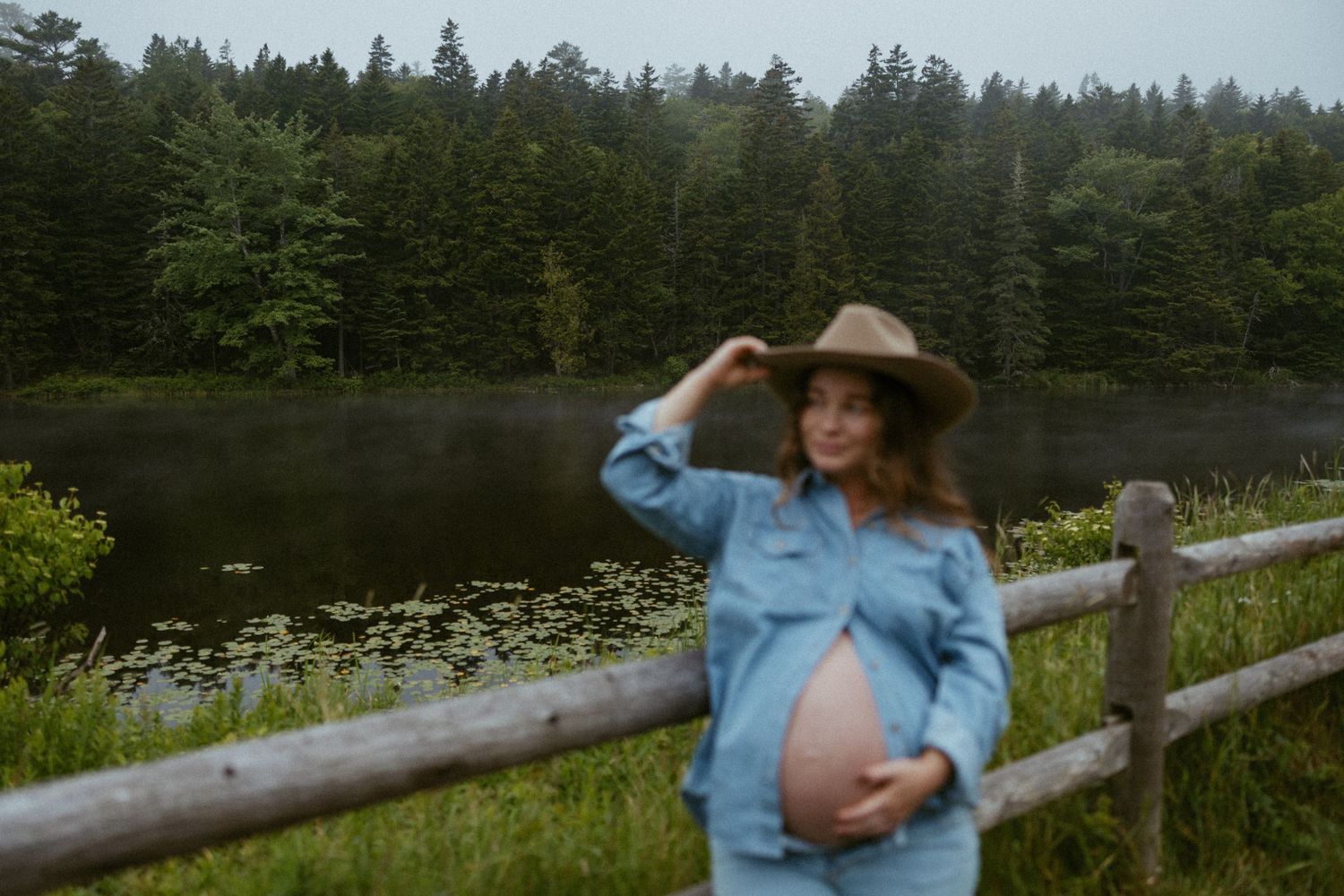 Pregnant woman in blue shirt and cowboy hat poses by rustic fence near misty lake surrounded by forest.