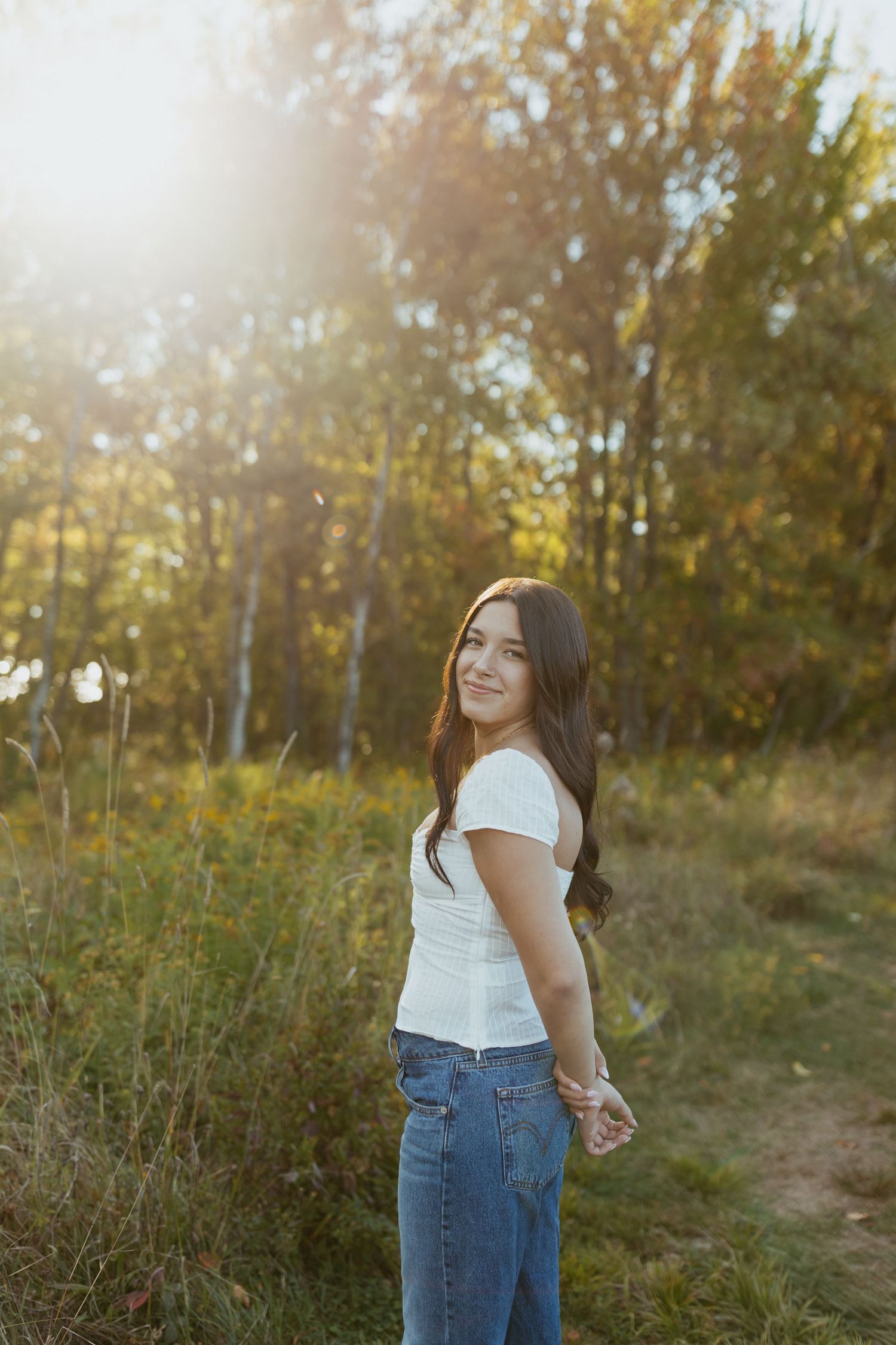 Young brunette woman in white top and jeans smiling over shoulder in sunlit autumn forest.