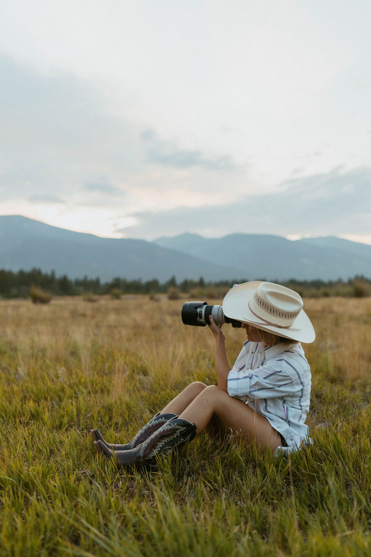 Woman in cowboy hat and boots sits in a field photographing mountain landscape at dusk.
