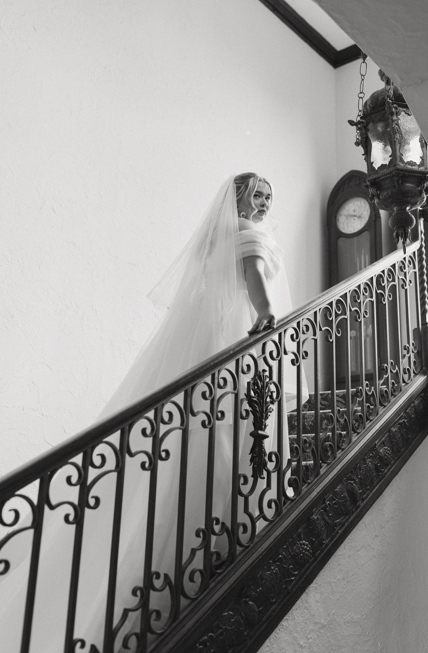 Bride in white gown and veil stands on ornate iron staircase beside vintage clock, black and white wedding photo.