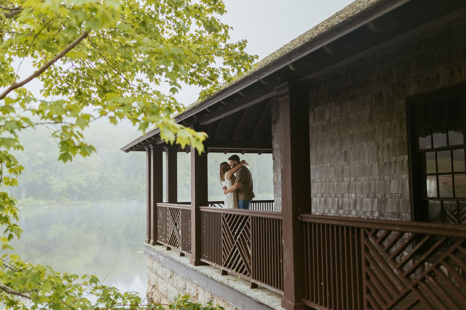 Couple sharing a romantic moment on a rustic wooden porch overlooking a misty pond surrounded by lush green trees.