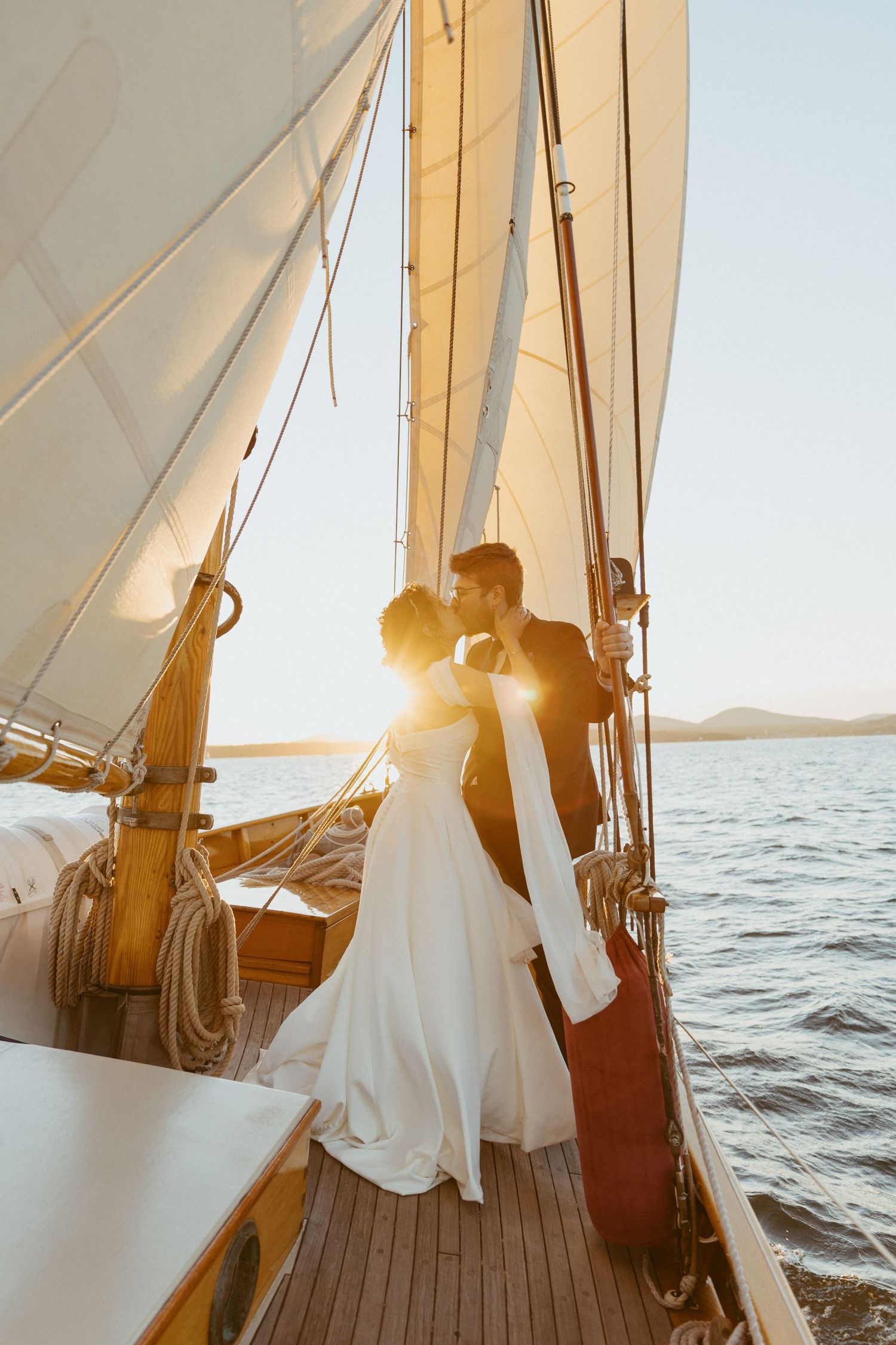 Bride and groom sharing a kiss on sailboat deck surrounded by white sails at sunset.