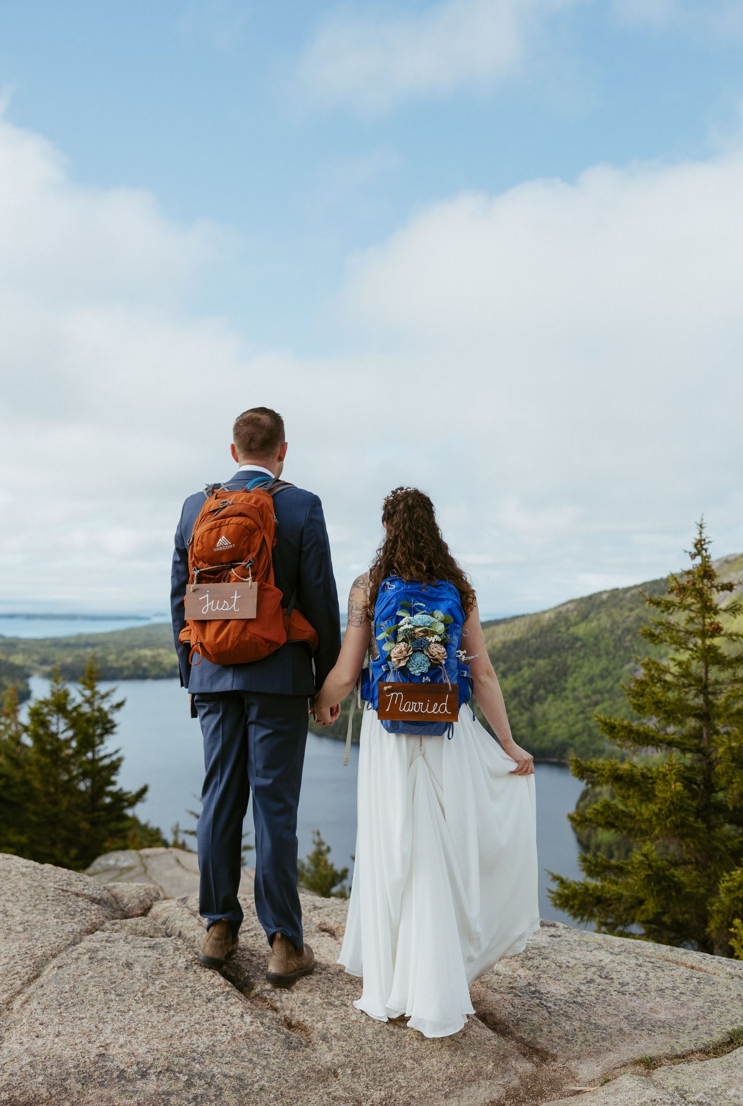 Newlywed couple with 'Just Married' backpacks standing on rocky overlook with scenic lake and forest views.