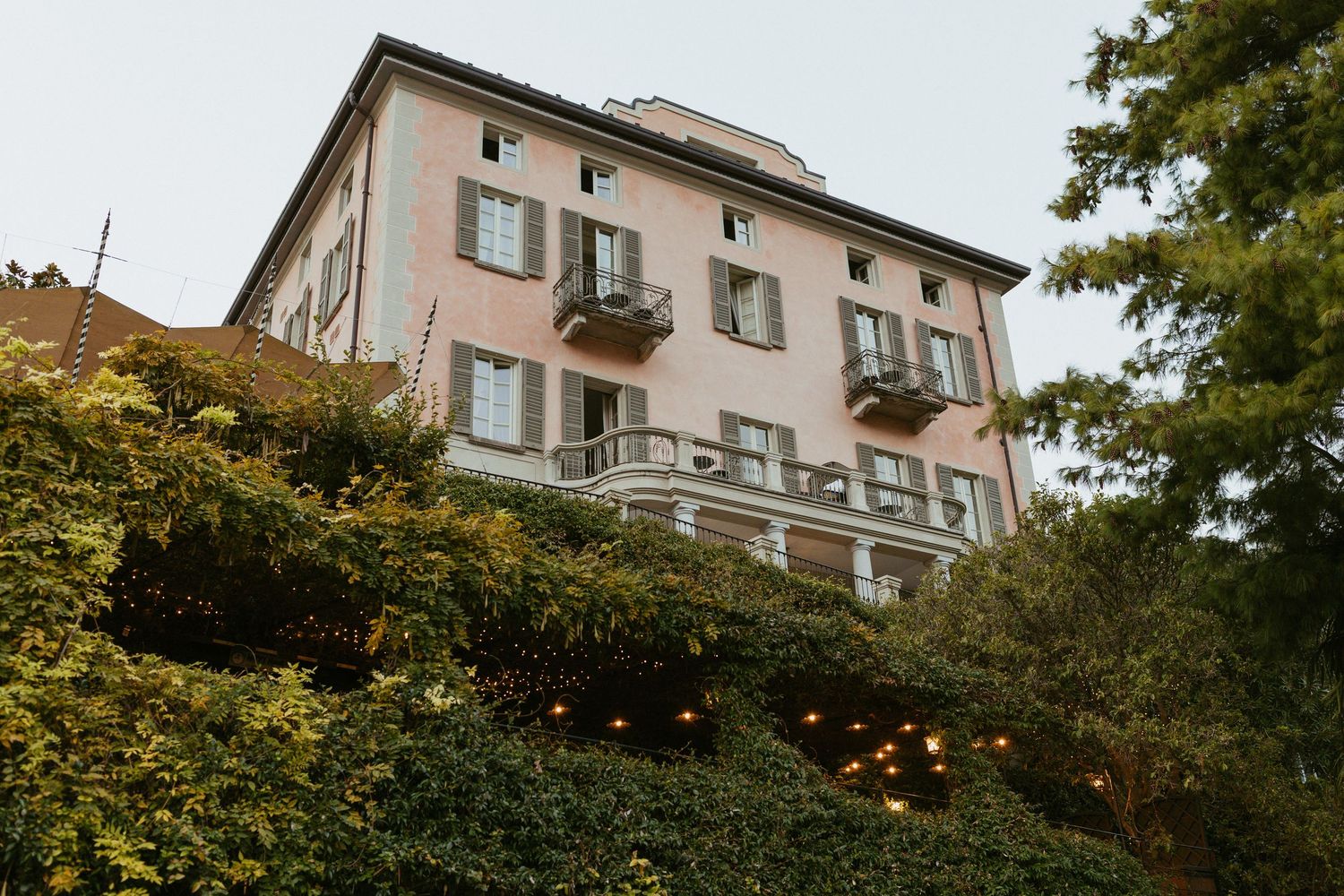 Pink Italian villa with green ivy, balconies, and warm lights nestled among lush trees on a hillside.