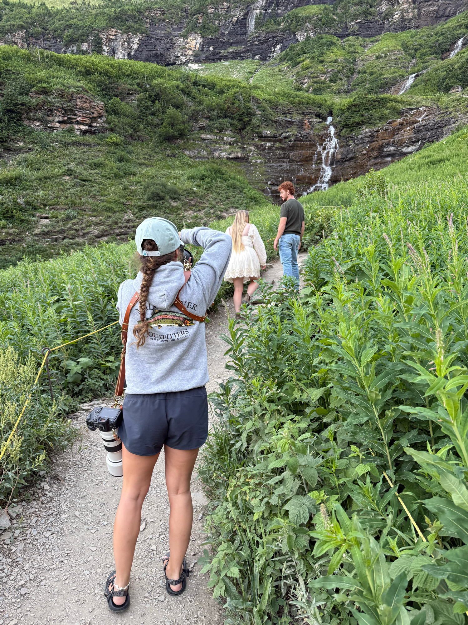Hikers walking a lush green mountain trail toward a waterfall, with a woman in foreground carrying a camera.