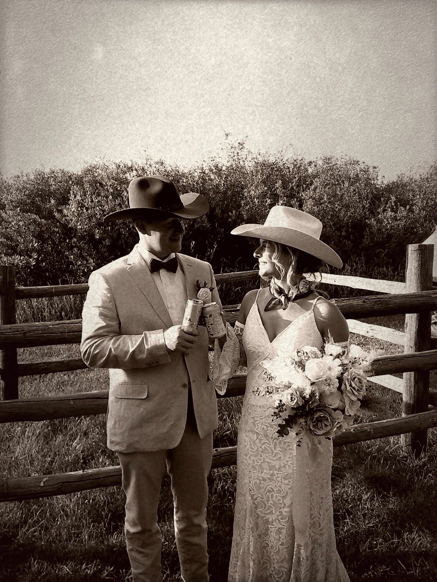 Couple in cowboy hats pose for rustic western wedding photo by wooden fence in black and white.