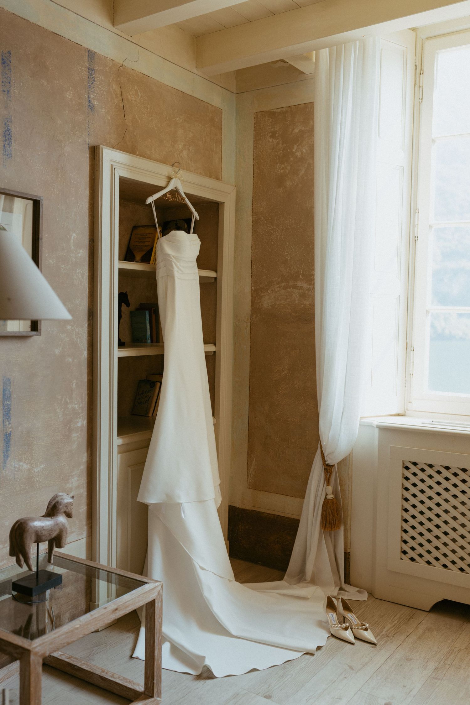 Elegant white wedding dress hanging on a mirror in a rustic Italian villa room with natural light.