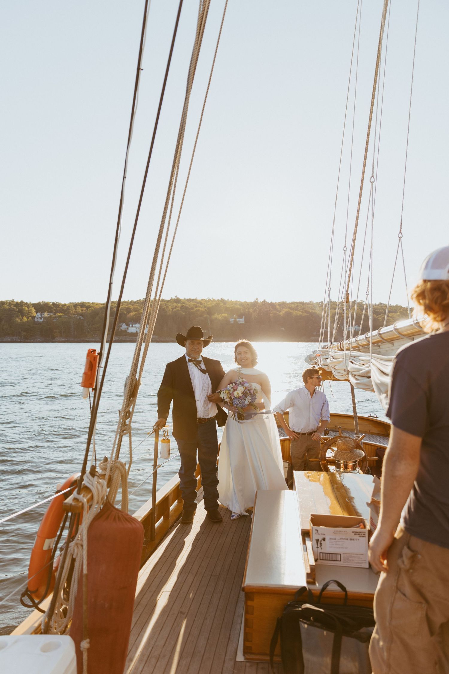 Wedding couple standing on sailboat deck at sunset with guests and bouquet.