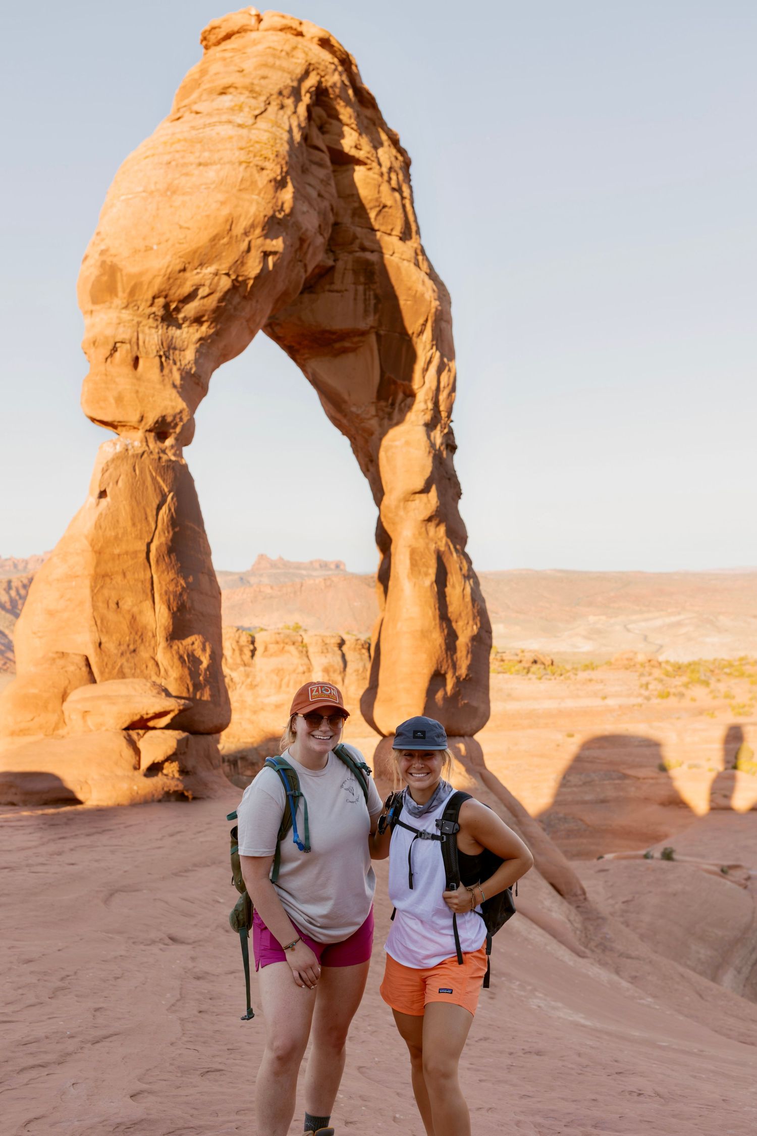 Two women hikers pose at Delicate Arch in Arches National Park at golden hour.