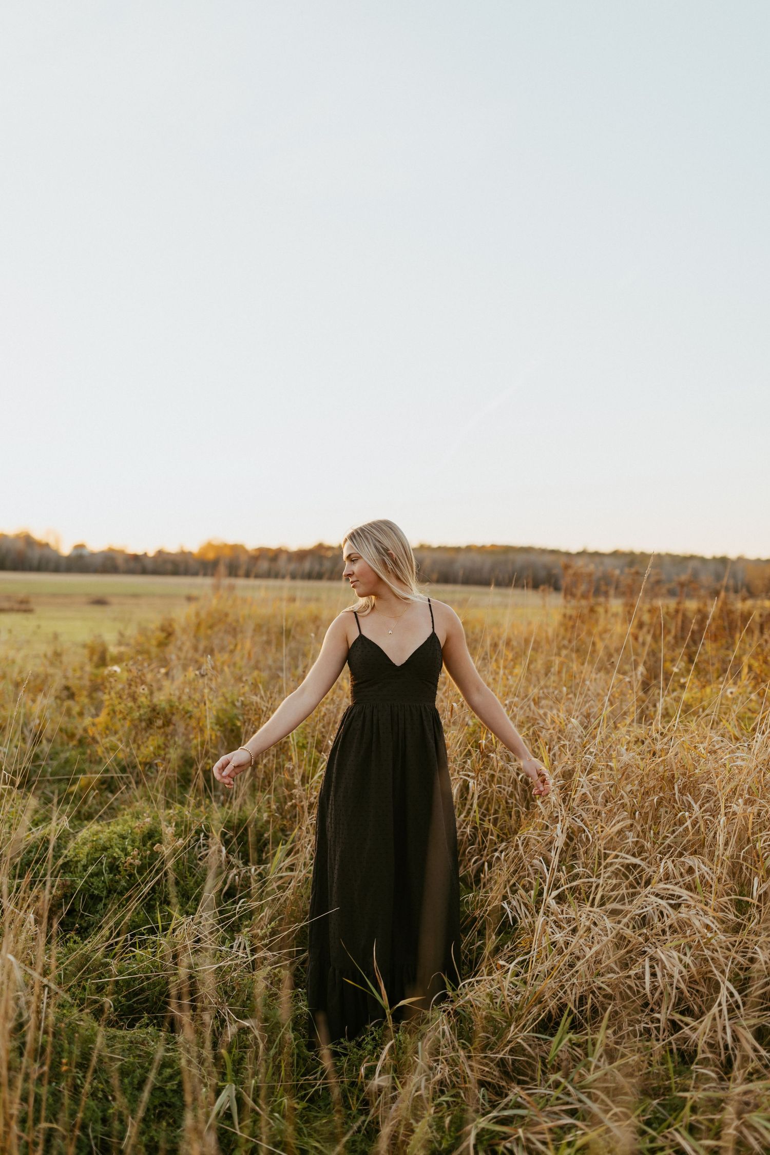 Blonde woman in black maxi dress walking through golden field at sunset.