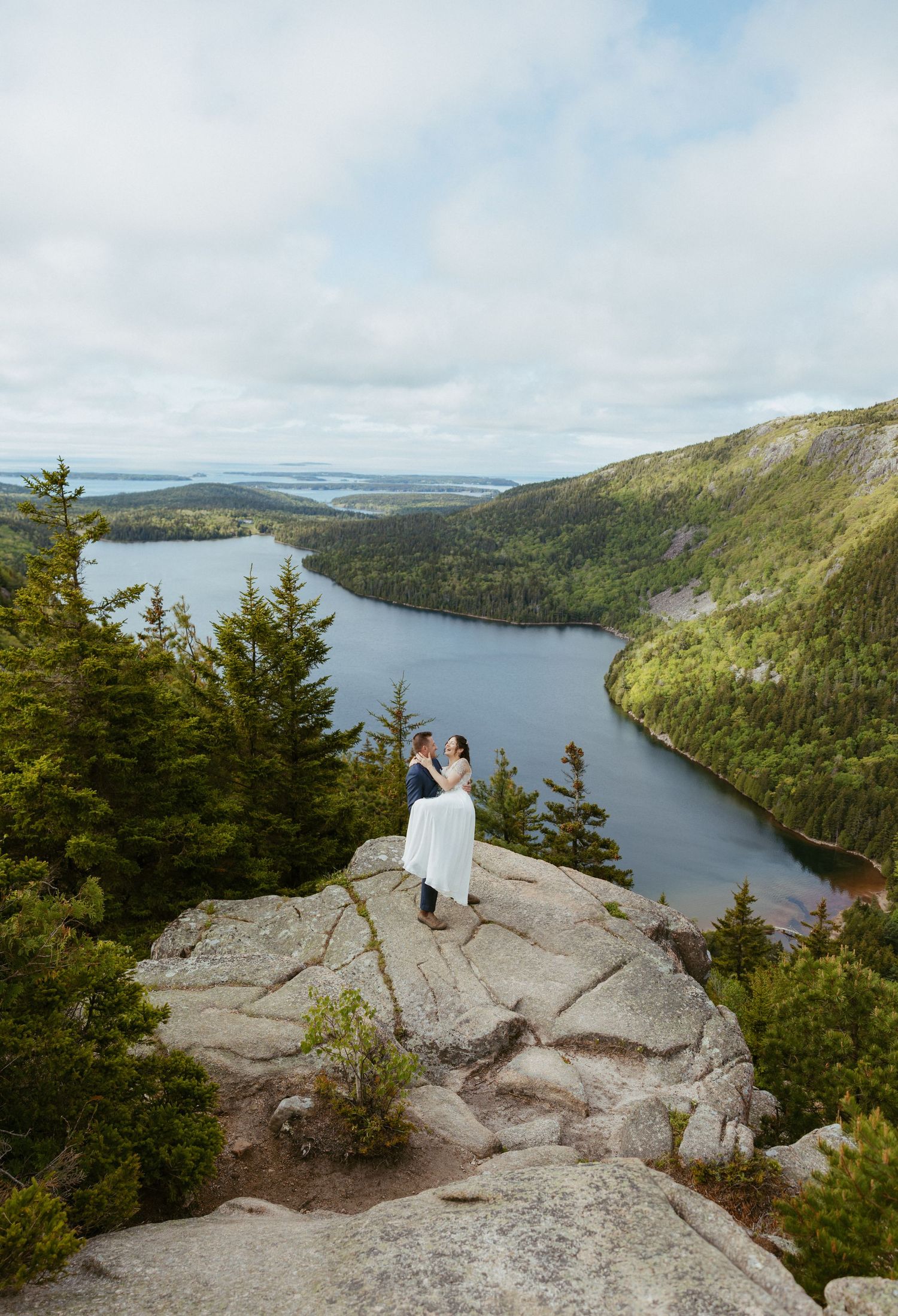 Couple embraces on rocky summit overlooking Jordan Pond in Acadia National Park, Maine, surrounded by lush forest.