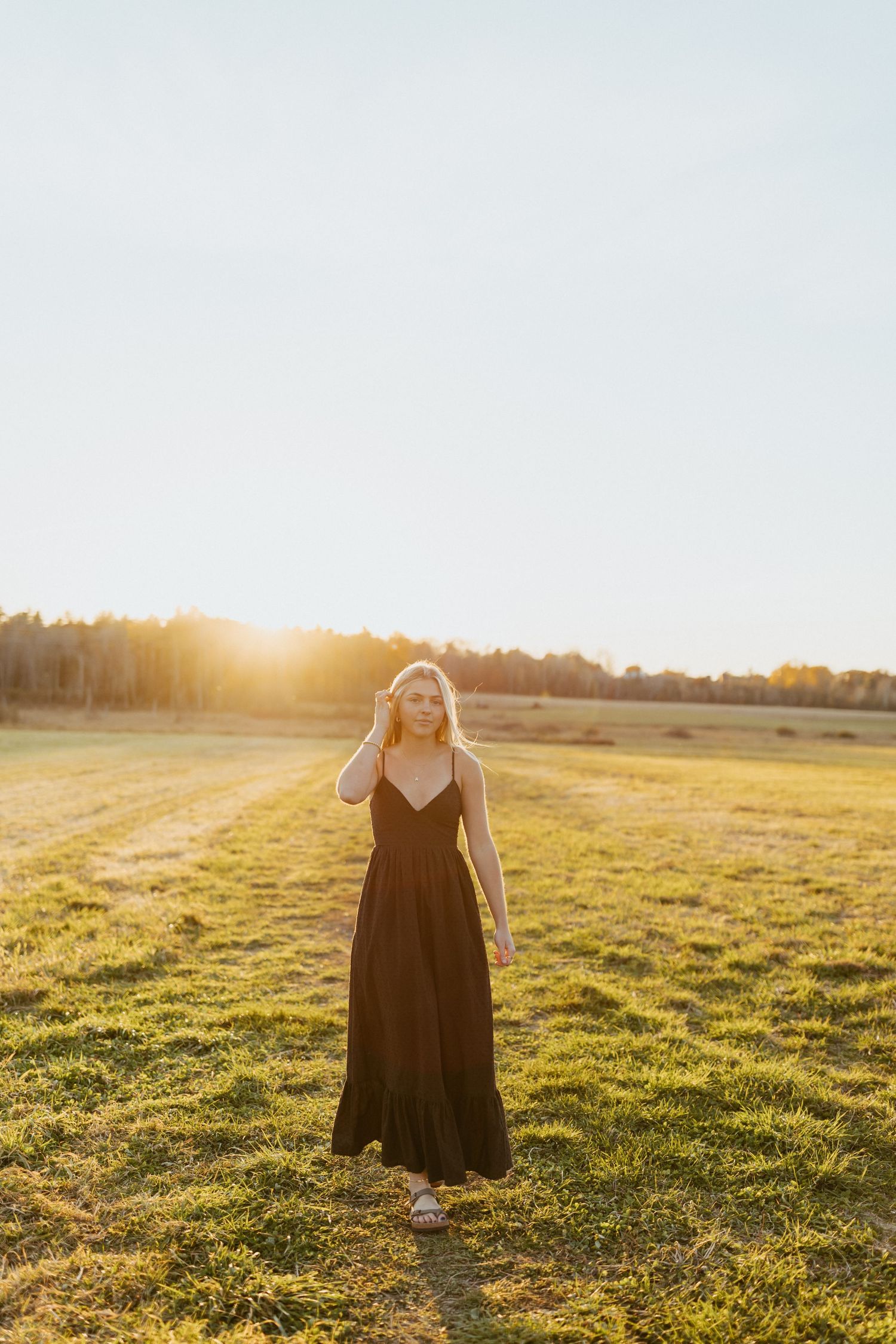 Woman in black maxi dress standing in sunlit field at golden hour with trees in background.