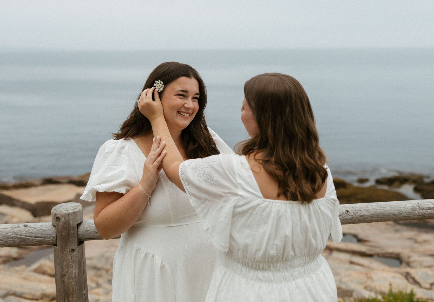 Two women in white dresses smile together near a rocky ocean overlook on a cloudy day.