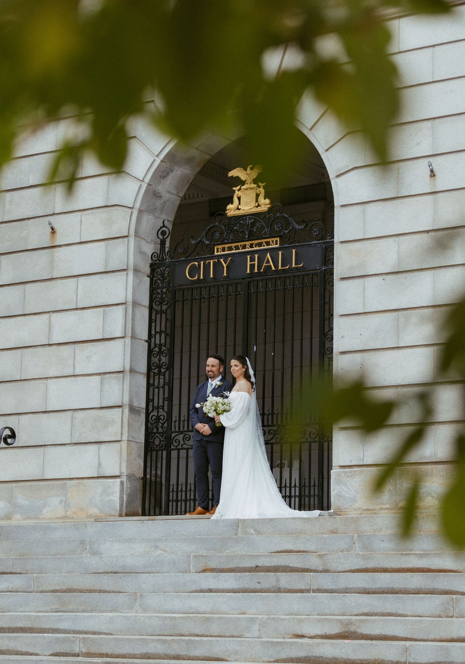 Bride and groom pose together on steps beneath ornate City Hall iron gates and archway.