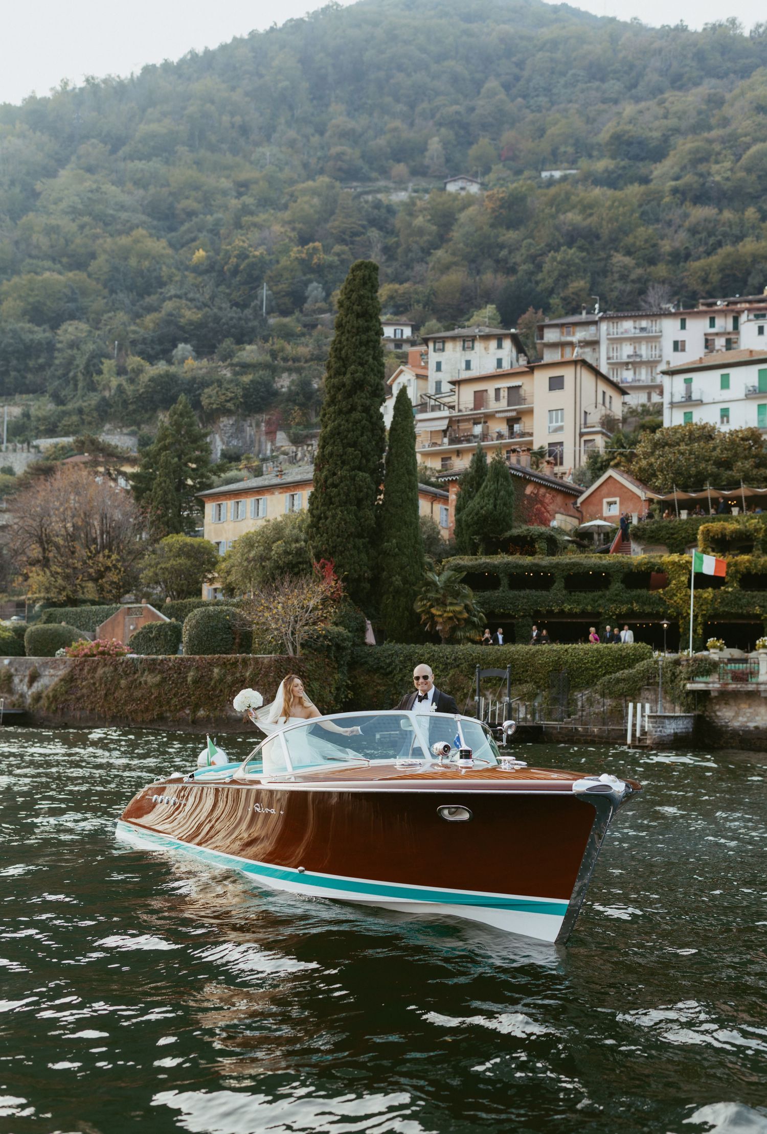 A classic wooden speedboat glides across Lake Como with hillside Italian village buildings in the background.