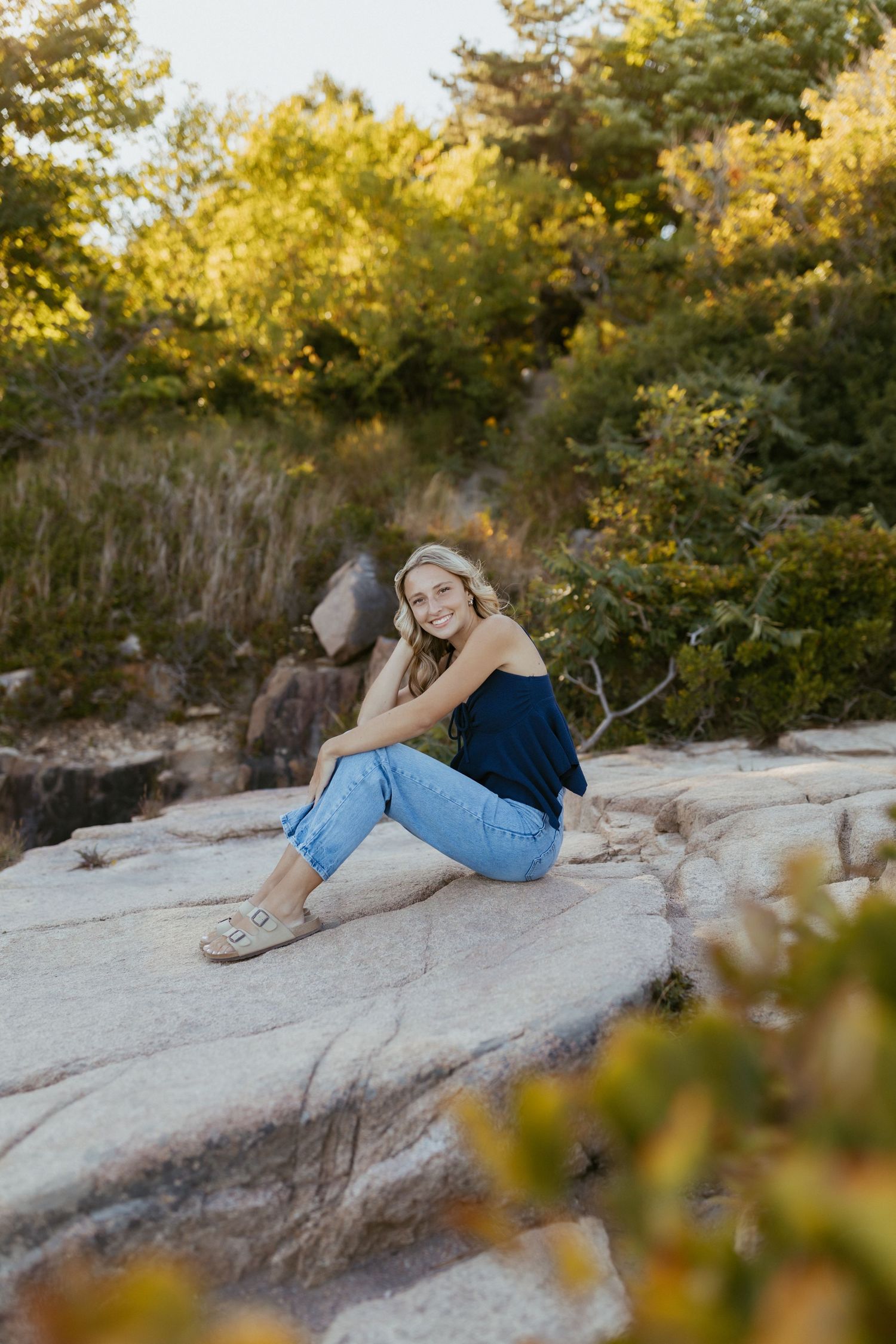 Smiling blonde woman sitting on rocky surface wearing navy tank and jeans with sandals outdoors.