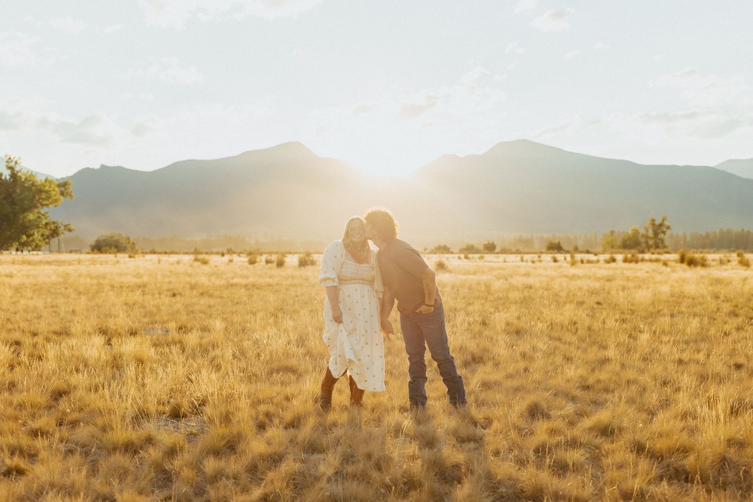 Couple sharing a romantic kiss in a golden field with Montana mountain backdrop at sunset.