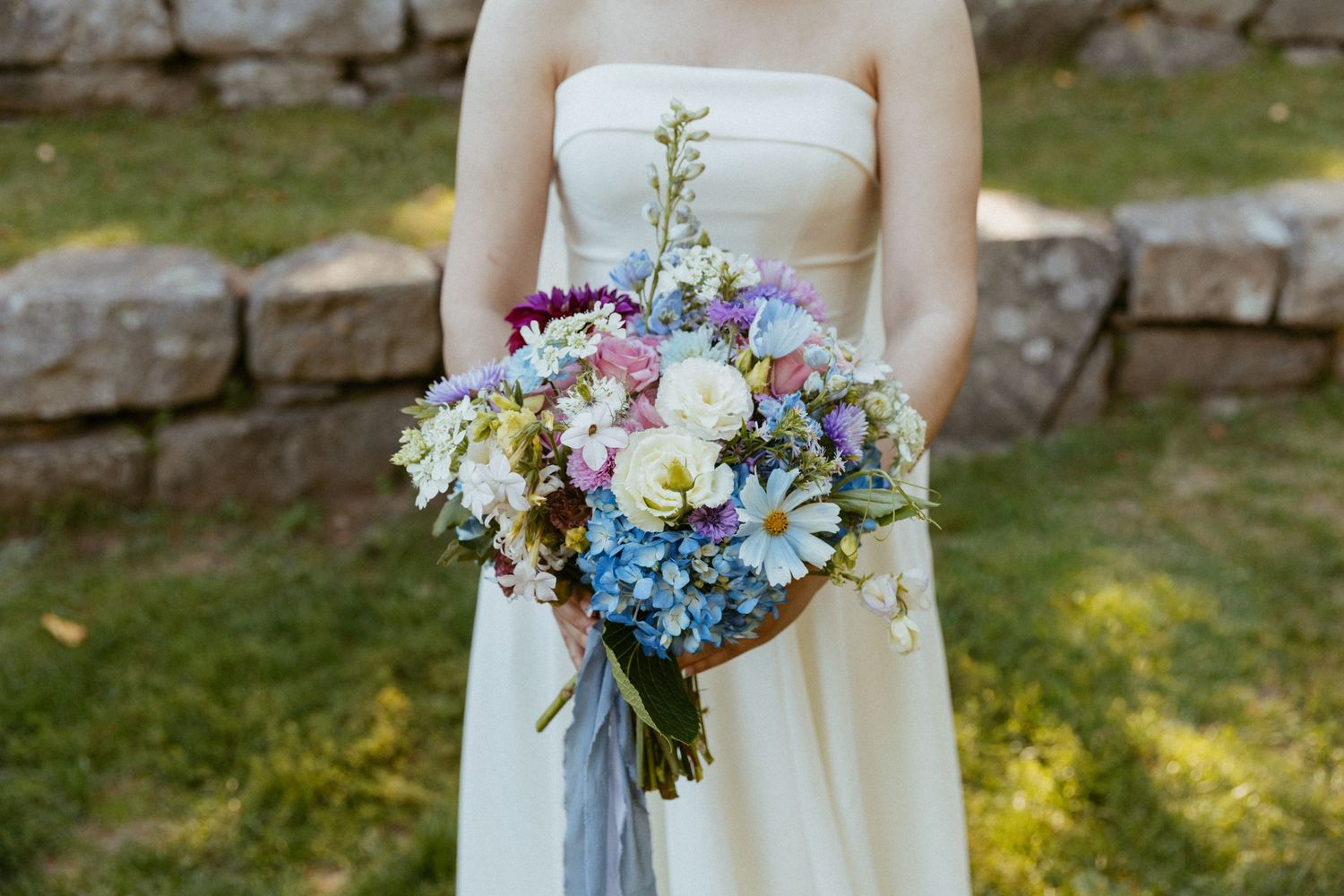 Bride holds a colorful wildflower bouquet with blue, white, and pink blooms in soft light.