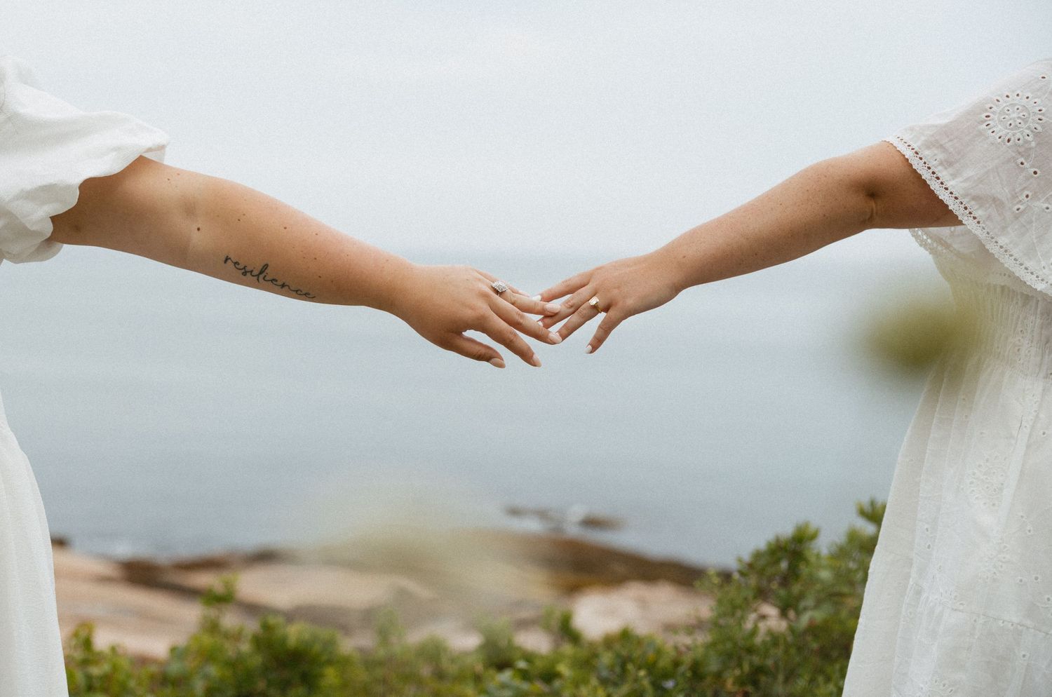 Two women in white dresses gently touching fingertips outdoors near a rocky shoreline.