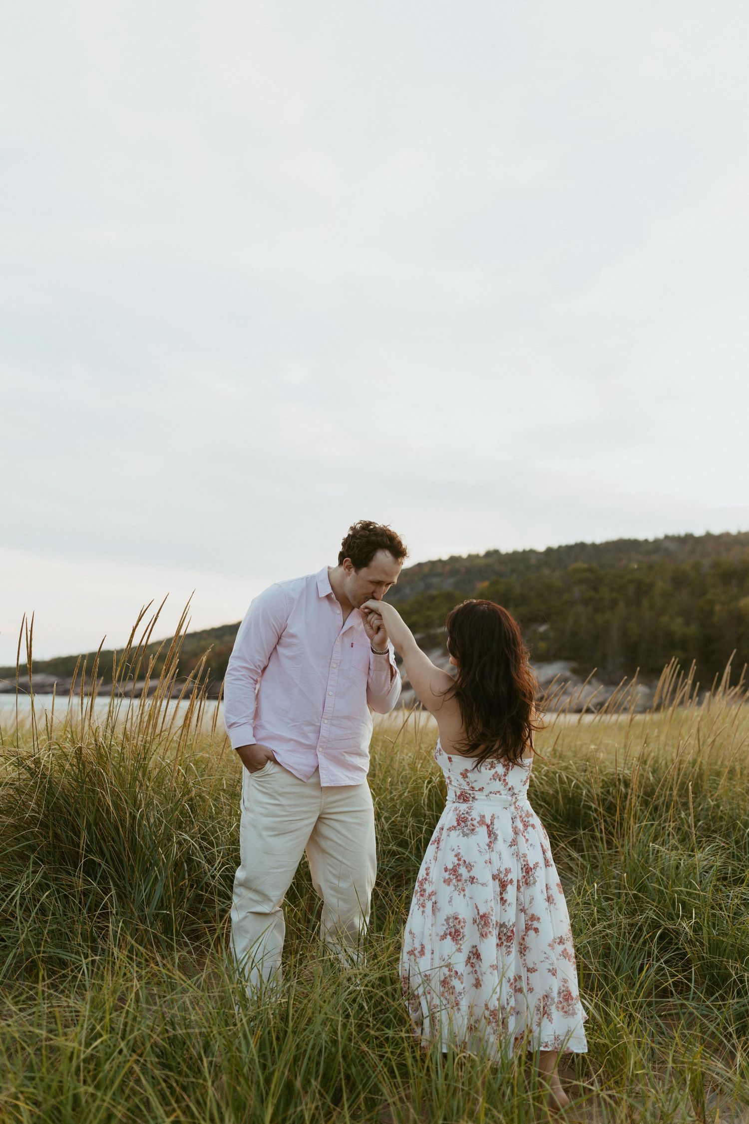 Romantic couple sharing tender moment in coastal dunes with ocean and hills behind.