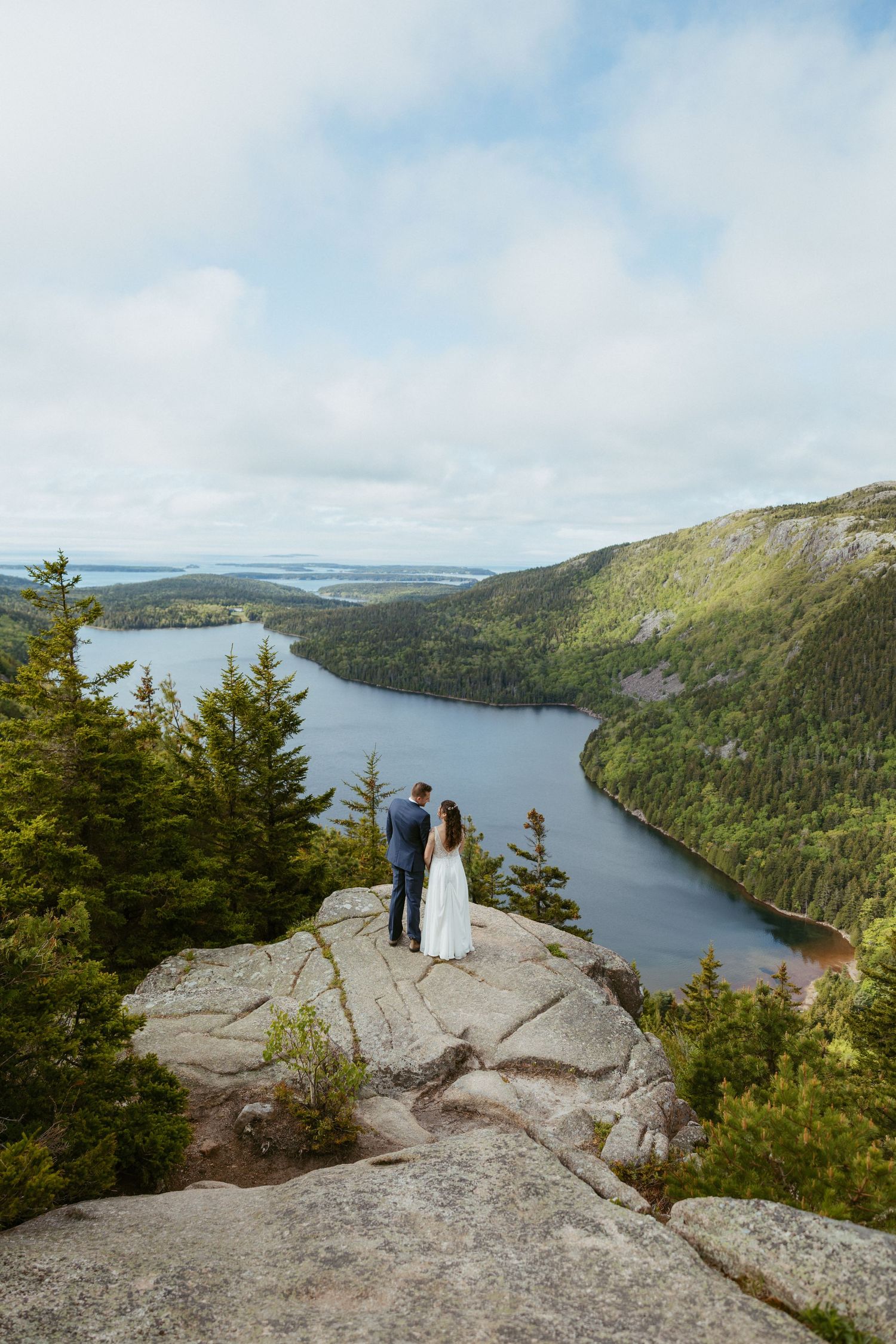 Couple in wedding attire standing on rocky overlook gazing at scenic lake and mountain views on their elopement day.