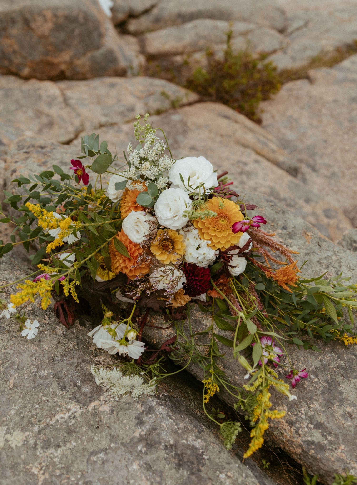 Wildflower wedding bouquet with white, orange, and yellow blooms resting on rustic stone rocks outdoors.