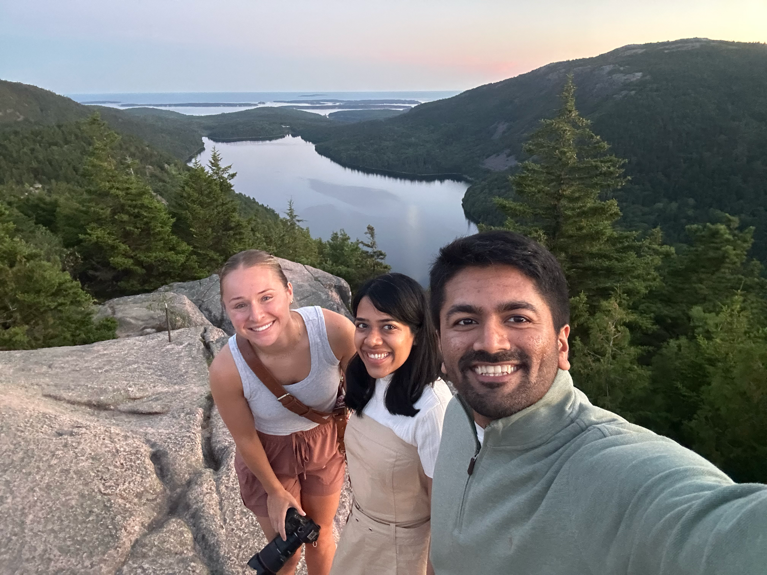 Three hikers smile for a selfie on a rocky summit with a scenic lake and forested mountains at sunset.
