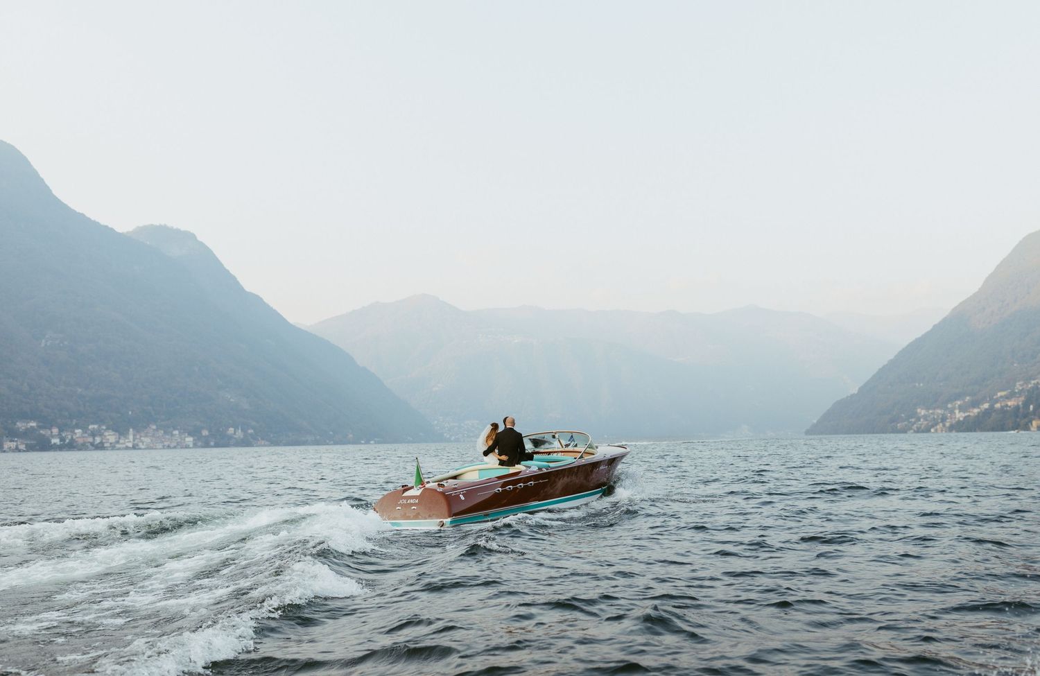 A classic wooden speedboat cruises on Lake Como with misty mountains in the background.